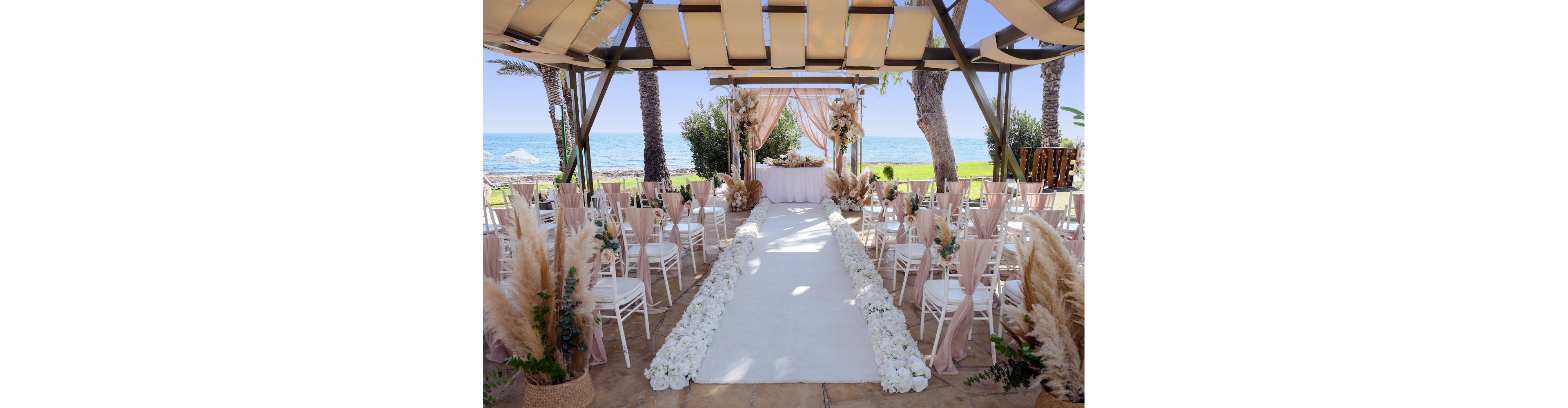 An outdoor wedding ceremony is arranged under a pergola overlooking the sea. A white aisle runner is lined with lush white flowers leading to a decorated altar with draped fabric and floral arrangements. Rows of white chairs with soft blush covers and greenery flank the aisle, creating a symmetrical, romantic setting. Pampas grass, palm trees, and the blue ocean in the background add a natural, coastal atmosphere.