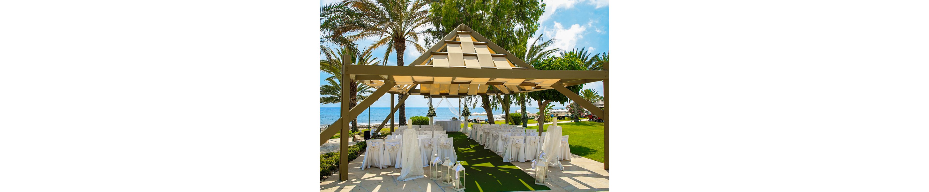 An elegant outdoor wedding ceremony area is arranged beneath a modern open pavilion near the sea. White-covered chairs and tables line a green aisle runner, leading toward the ocean view framed by palm trees. Decorative lanterns and bows add a romantic touch to the bright, sunlit setting. The scene combines lush greenery, blue water, and clear skies for a serene coastal celebration atmosphere.