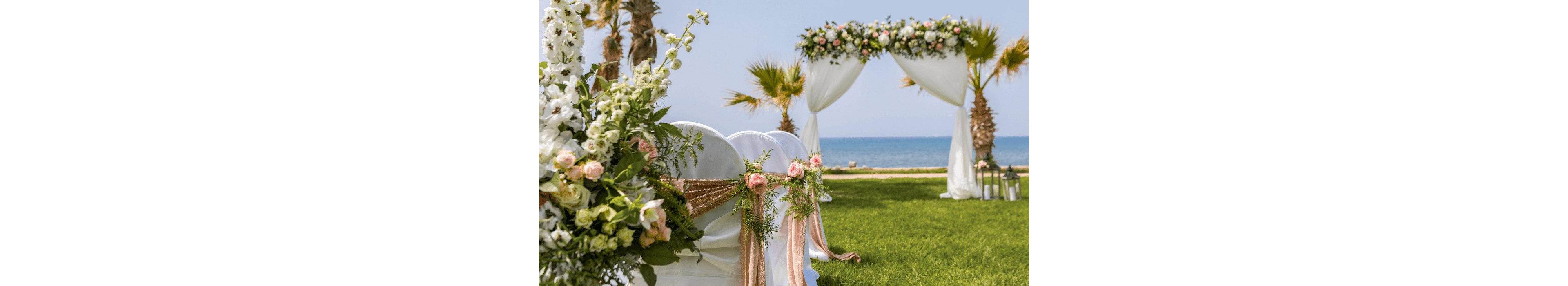 An outdoor wedding ceremony setup is arranged on a lush green lawn by the sea. White chairs are decorated with soft pink and white flowers and draped fabric. A floral arch with white curtains stands in the background, framing the ocean and palm trees. The bright daylight and pastel decorations create a romantic, elegant atmosphere.