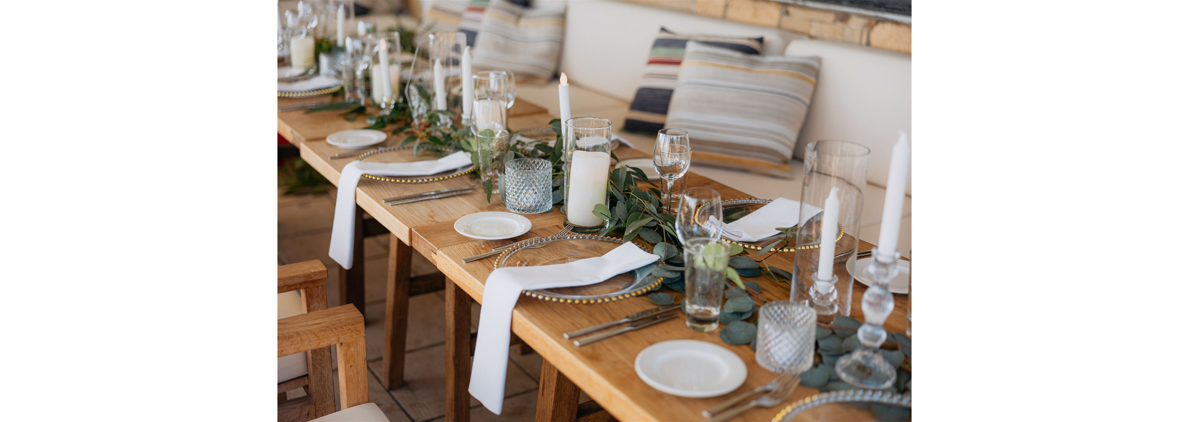 A long wooden table is set for a formal meal with white plates, glassware, and silver cutlery. Tall white taper candles and glass candle holders run down the center alongside a lush green foliage garland. White cloth napkins hang over the edge of each place setting, adding a refined touch. Cushioned bench seating with striped pillows creates a relaxed yet sophisticated atmosphere suitable for a celebration or intimate gathering.