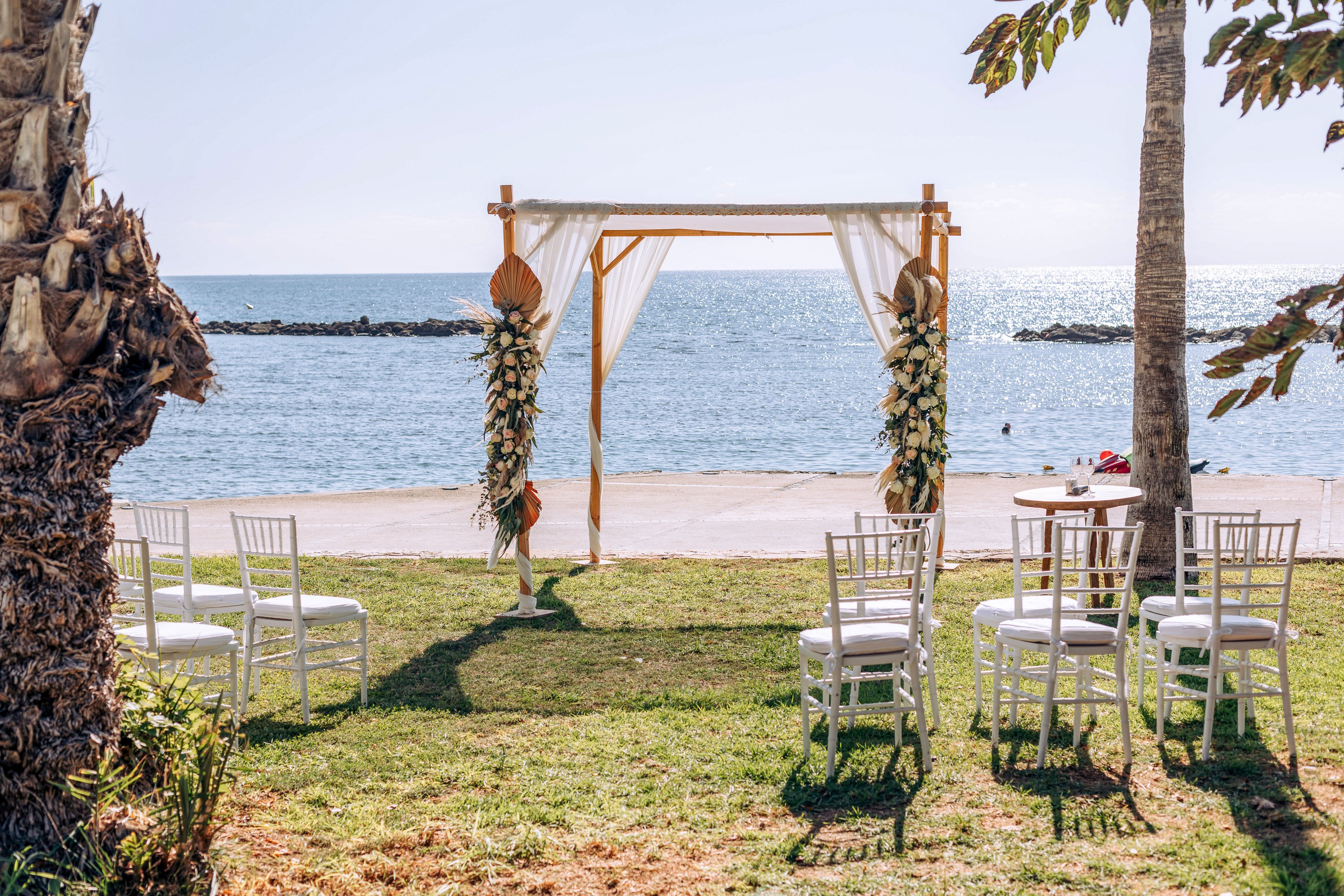 A wooden wedding arch decorated with white drapery and floral arrangements stands on a grassy lawn by the sea. White chairs are arranged in small rows facing the arch, creating an intimate ceremony setting. Palm trees frame the scene, and calm blue water stretches out to the horizon under a clear sky. The overall atmosphere is bright, romantic, and serene, ideal for a beachside celebration.