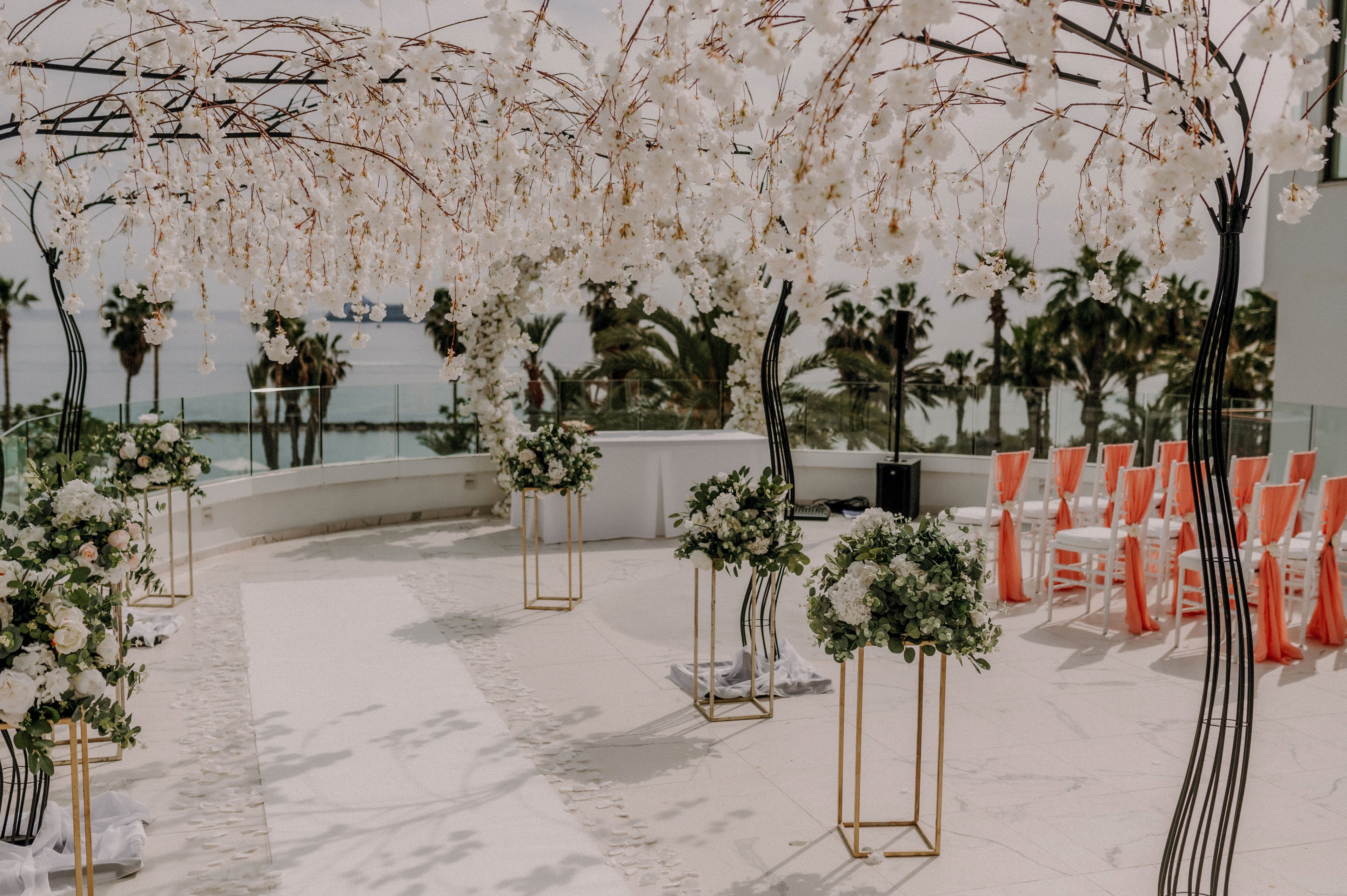 An outdoor wedding ceremony setup features a white aisle framed by tall floral arrangements and delicate hanging blossoms. Orange chairs are neatly arranged to one side, facing a simple altar area with a view of the ocean. Palm trees and a glass railing in the background suggest a coastal venue with a modern, open-air design. The overall scene feels romantic, airy, and sophisticated, emphasizing soft white tones with bright orange accents.