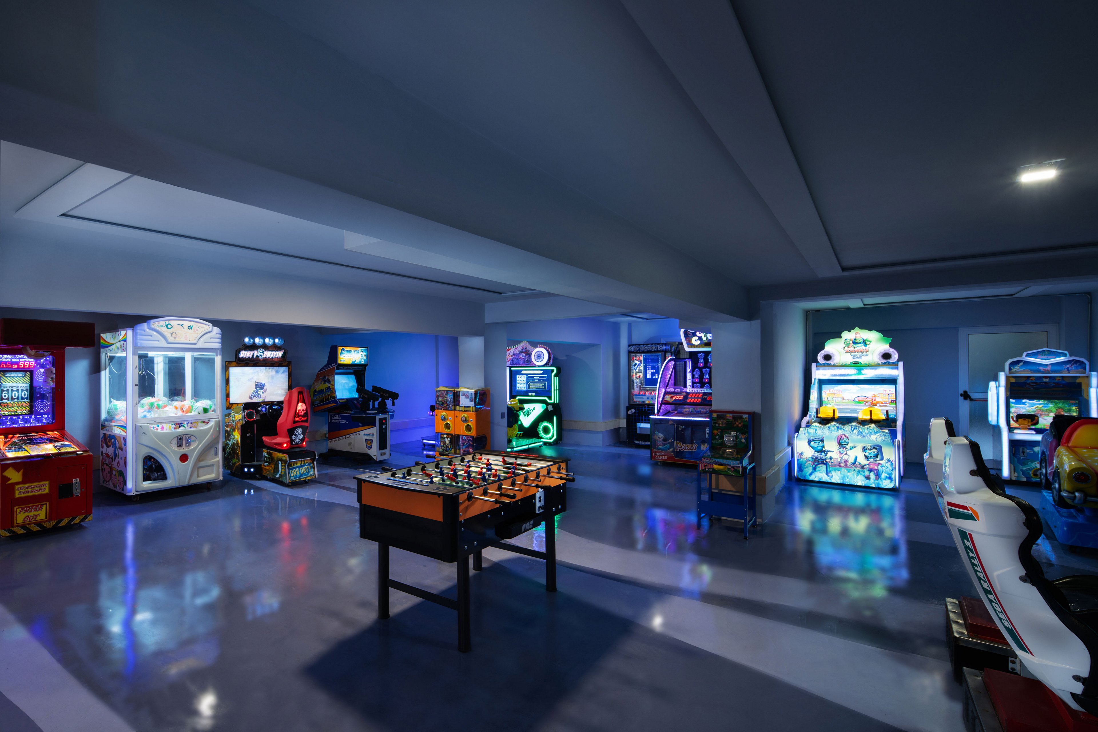 A spacious arcade room featuring various gaming machines and activities. The setting includes foosball tables, racing simulators, and classic arcade games illuminated by colorful neon lights. The polished floor reflects the vibrant glow, creating an energetic atmosphere. The room is designed for entertainment and leisure, appealing to all age groups.