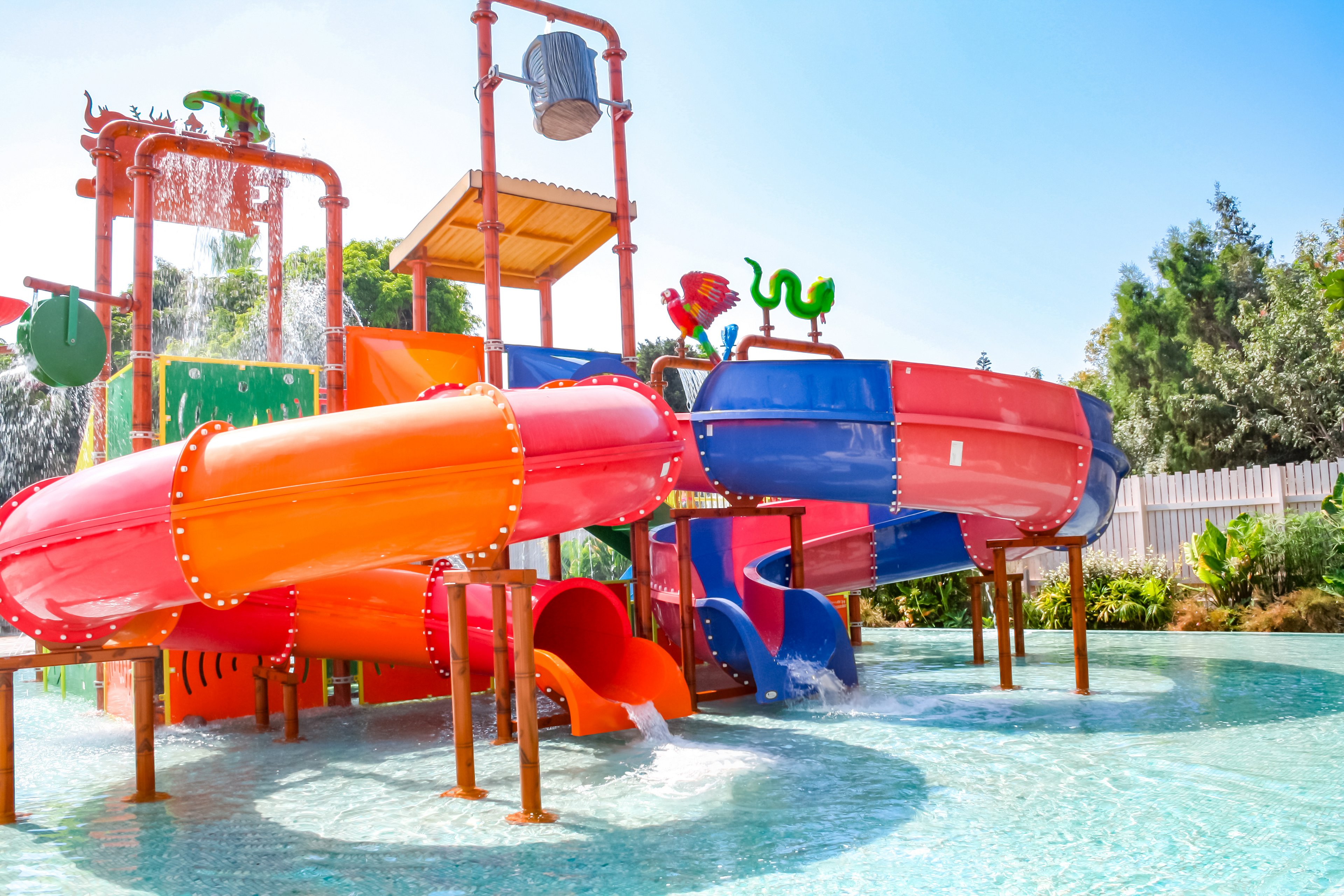 A vibrant water park featuring colorful slides in orange, purple, and red. Children are enjoying the shallow water area under sunny skies. The setting includes playful structures and water features surrounded by greenery and fencing.