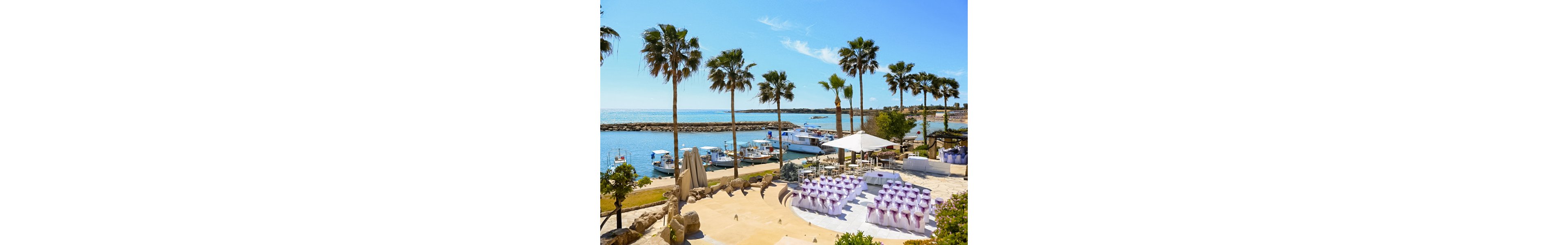 An outdoor wedding ceremony setup overlooks a sunny marina with several docked boats. Rows of white chairs decorated with purple bows are arranged facing a small altar area near the water. Tall palm trees frame the scene under a clear blue sky, creating a bright and festive atmosphere. The setting suggests a coastal resort or beachside venue prepared for a special event.