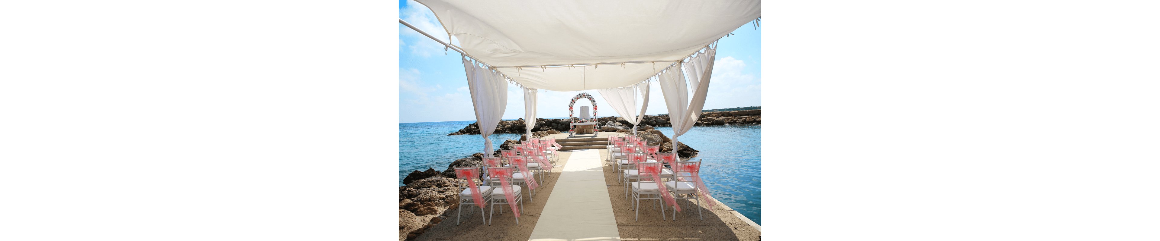 An outdoor wedding ceremony setup is arranged beneath a white fabric canopy by the sea. Rows of white chairs with pink sashes line a central aisle leading to a small altar at the end of a stone pier. A floral arch frames the altar, with calm blue water and rocky shoreline in the background. The bright daylight and soft pastel decorations create a romantic, serene atmosphere.