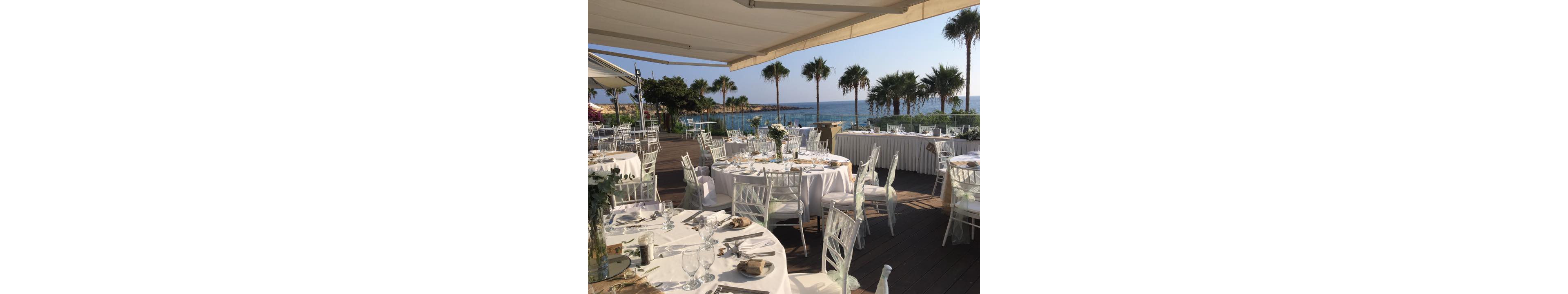 An elegant outdoor wedding reception is arranged on a wooden deck overlooking the sea. Round tables with white linens, glassware, and floral centerpieces are neatly set under large awnings. White chiavari chairs surround the tables, creating a refined and airy atmosphere. Palm trees, calm blue water, and clear skies enhance the serene coastal setting.