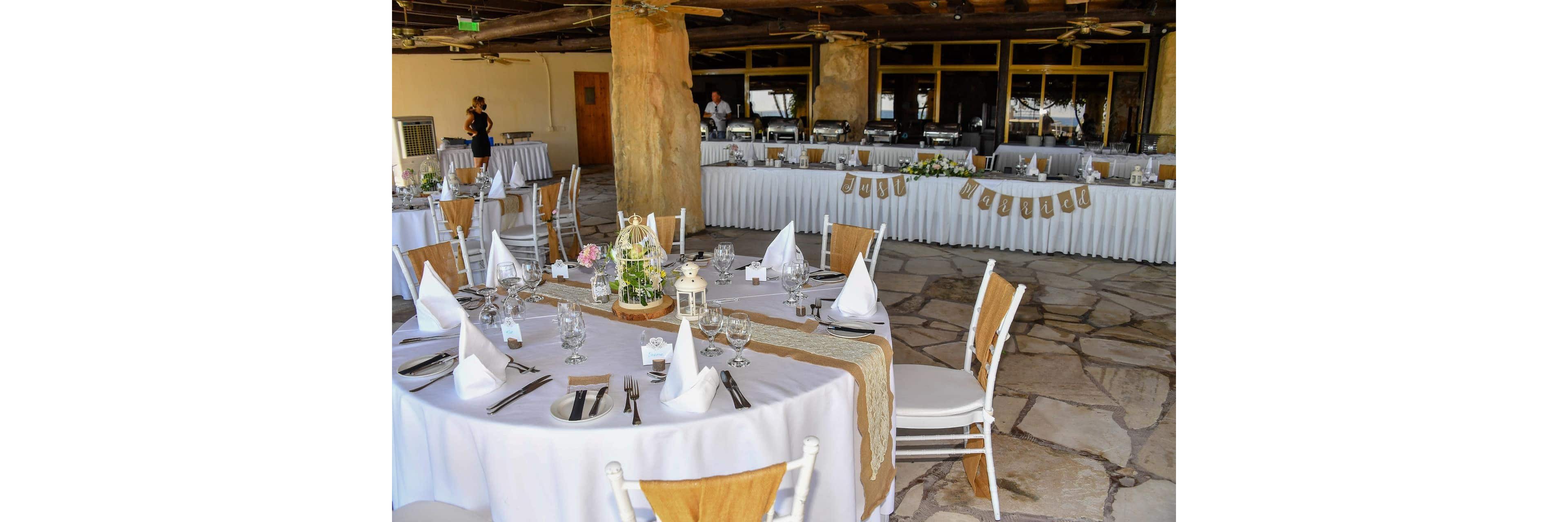 Round banquet tables are elegantly set for a rustic wedding reception in an open-air venue. White tablecloths, folded napkins, and simple floral centerpieces decorate the tables, complemented by burlap runners and wooden accents. A long buffet table is arranged along the back wall, ready for guests. The stone floor, wooden ceiling, and natural pillars create a warm, casual atmosphere. A few people stand in the background, adding a sense of anticipation before the event begins.