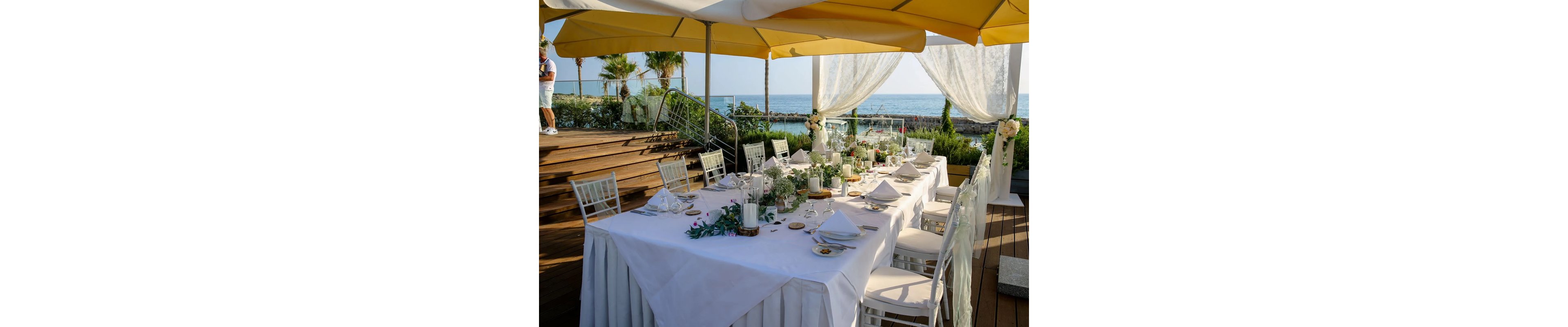 A long, white-clothed table is beautifully set for an outdoor event, likely a wedding reception. The scene is arranged on a wooden deck with white chairs, candles, floral centerpieces, and neatly folded napkins. Large yellow umbrellas and sheer white drapes frame the view of the ocean and rocky shoreline in the background. Lush greenery and palm trees add to the bright, summery atmosphere.