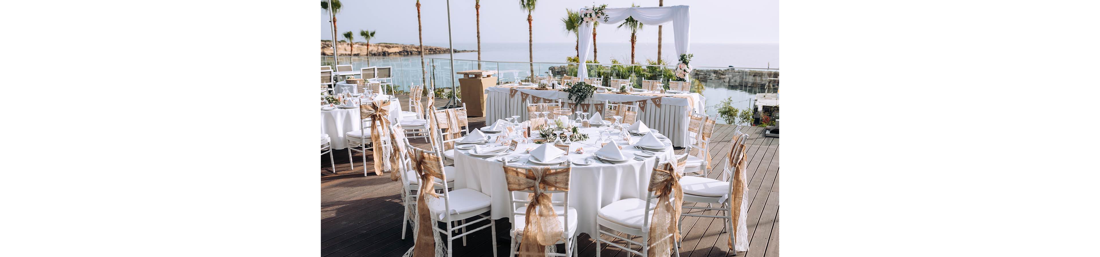An outdoor wedding reception is arranged on a wooden deck overlooking the sea. Round tables are covered with white linens and decorated with rustic burlap chair sashes and floral centerpieces. A white ceremonial arch and a long head table stand near the water, framed by palm trees. The scene feels bright and romantic, with soft natural light and a coastal backdrop.