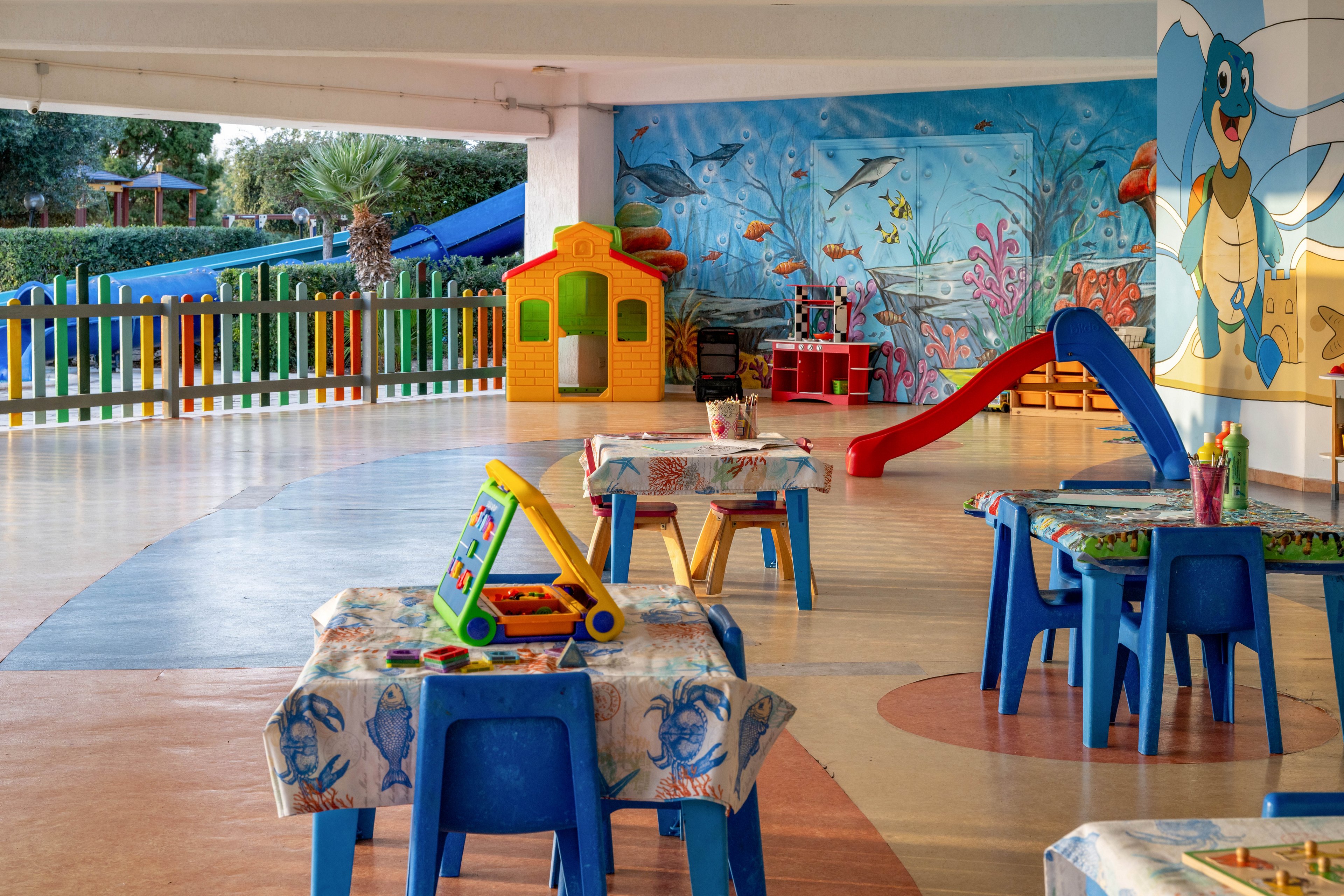 A bright covered play area features small tables and chairs arranged for children’s activities. In the background, a red slide and a yellow playhouse sit in front of a colorful ocean-themed mural with fish and sea plants. The tables are covered with patterned cloths and hold various toys and art supplies. A fenced outdoor area with trees and additional play structures is visible beyond the open side of the space.