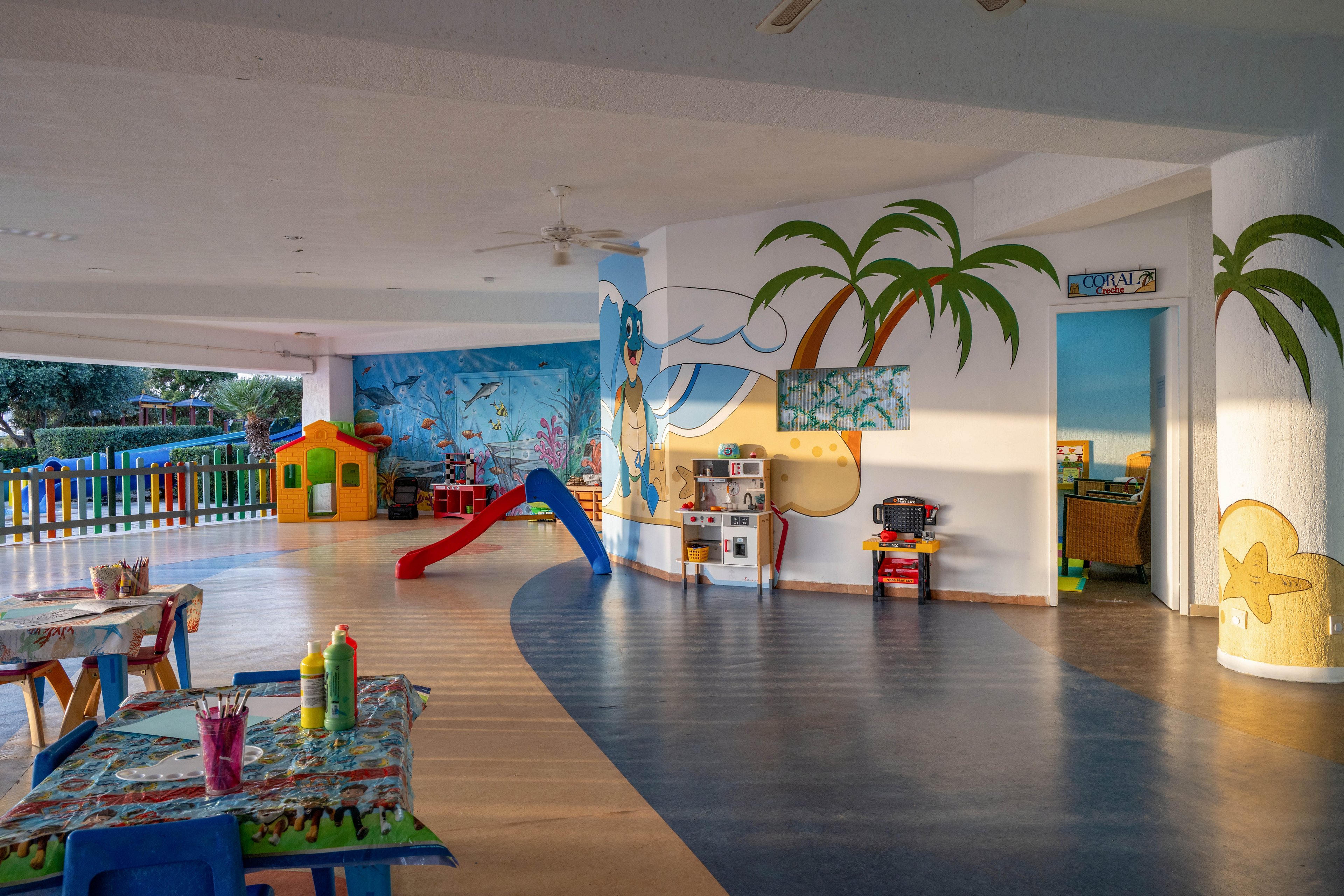A bright indoor playroom features a tropical-themed mural with palm trees and ocean scenes on the walls and columns. Small tables with art supplies, including paint bottles and brushes, are arranged near colorful chairs. A red slide, toy house, and various play stations line the curved floor, creating a fun and engaging environment for children. Natural light enters from the side, enhancing the cheerful and welcoming atmosphere.