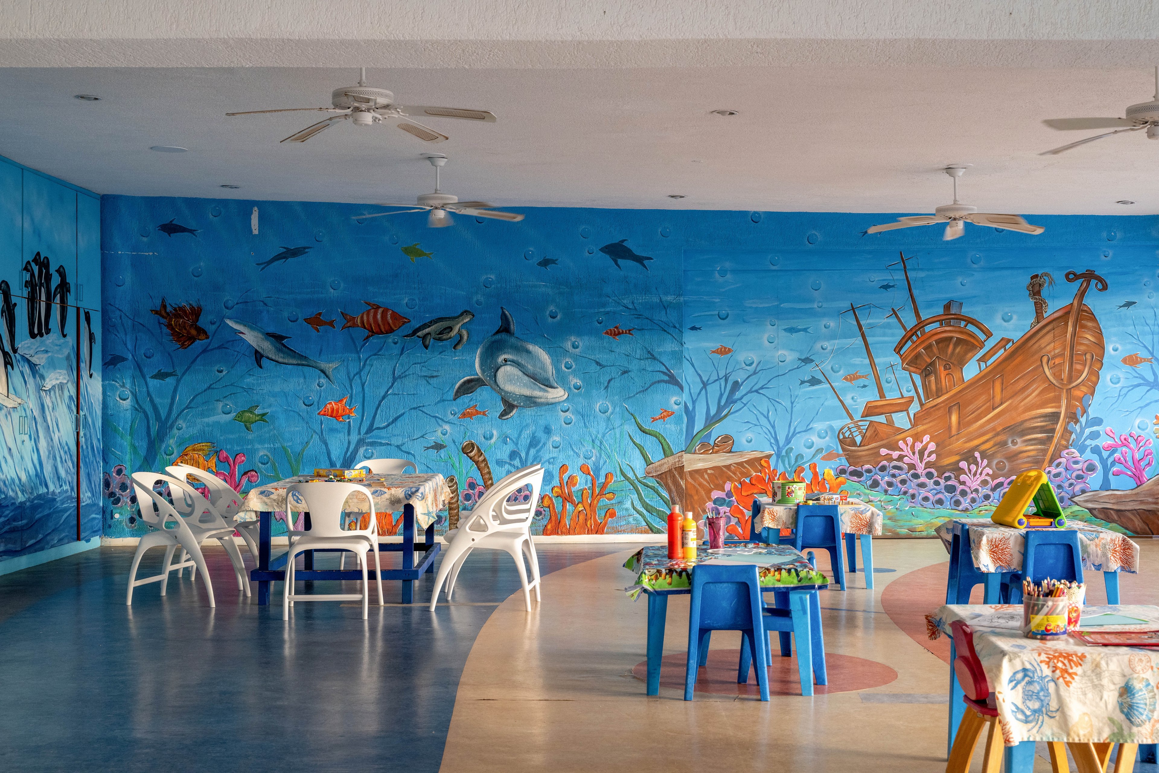 A bright children's art room features small tables and chairs arranged beneath a large underwater-themed wall mural. The mural depicts sea creatures, coral reefs, and a sunken wooden ship in vibrant blues and oranges. Art supplies and paint bottles sit on the tables, suggesting a creative activity space. Ceiling fans and open floor space contribute to a relaxed, playful environment.