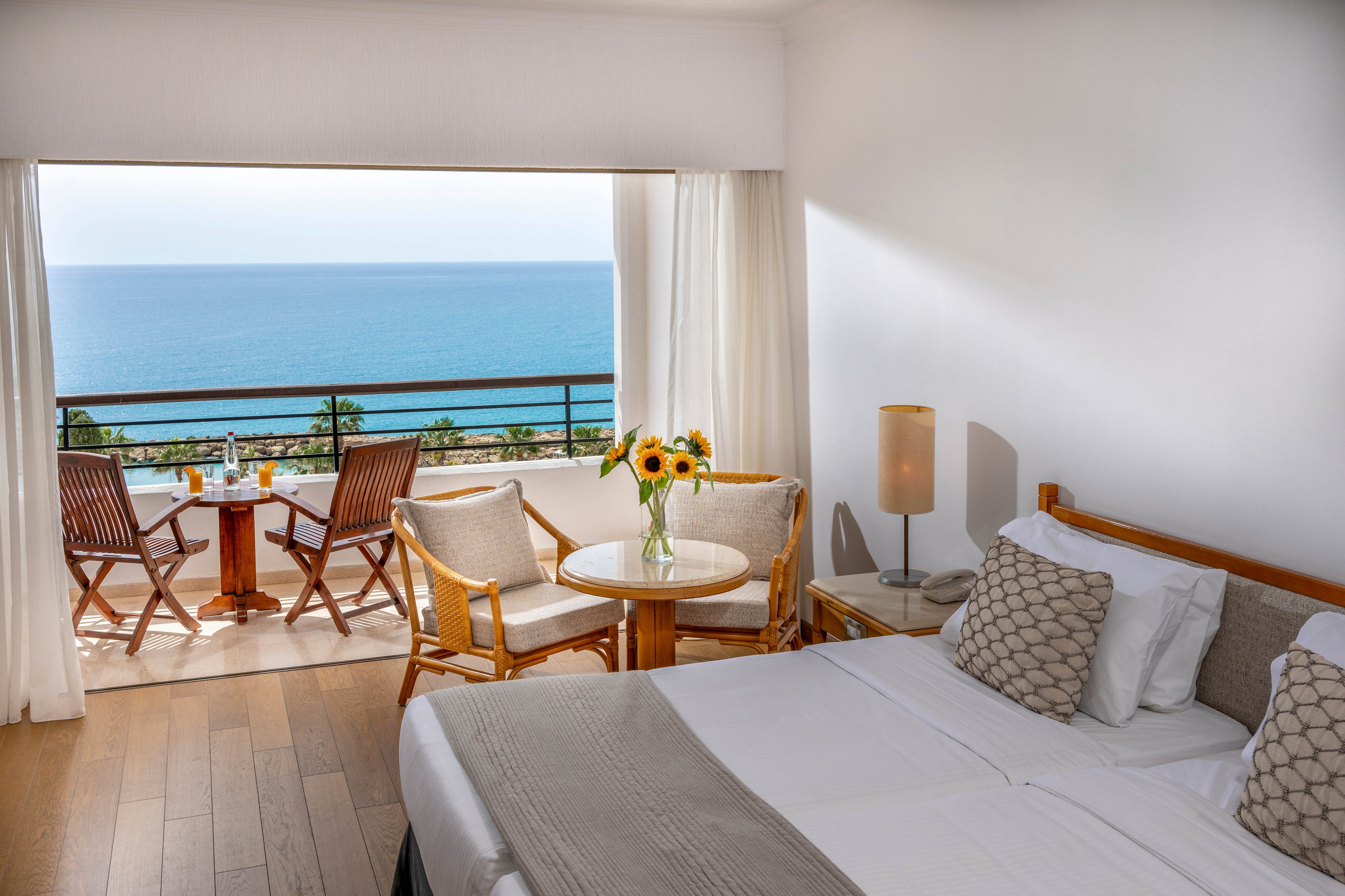 A modern hotel bedroom features a neatly made double bed with neutral-toned linens and patterned pillows. Large sliding doors open onto a balcony overlooking a calm, expansive ocean. The balcony is furnished with wooden chairs and a small round table set for two, including drinks and a vase of sunflowers. Warm natural light fills the room, highlighting the wooden floor, minimalist decor, and serene coastal setting.