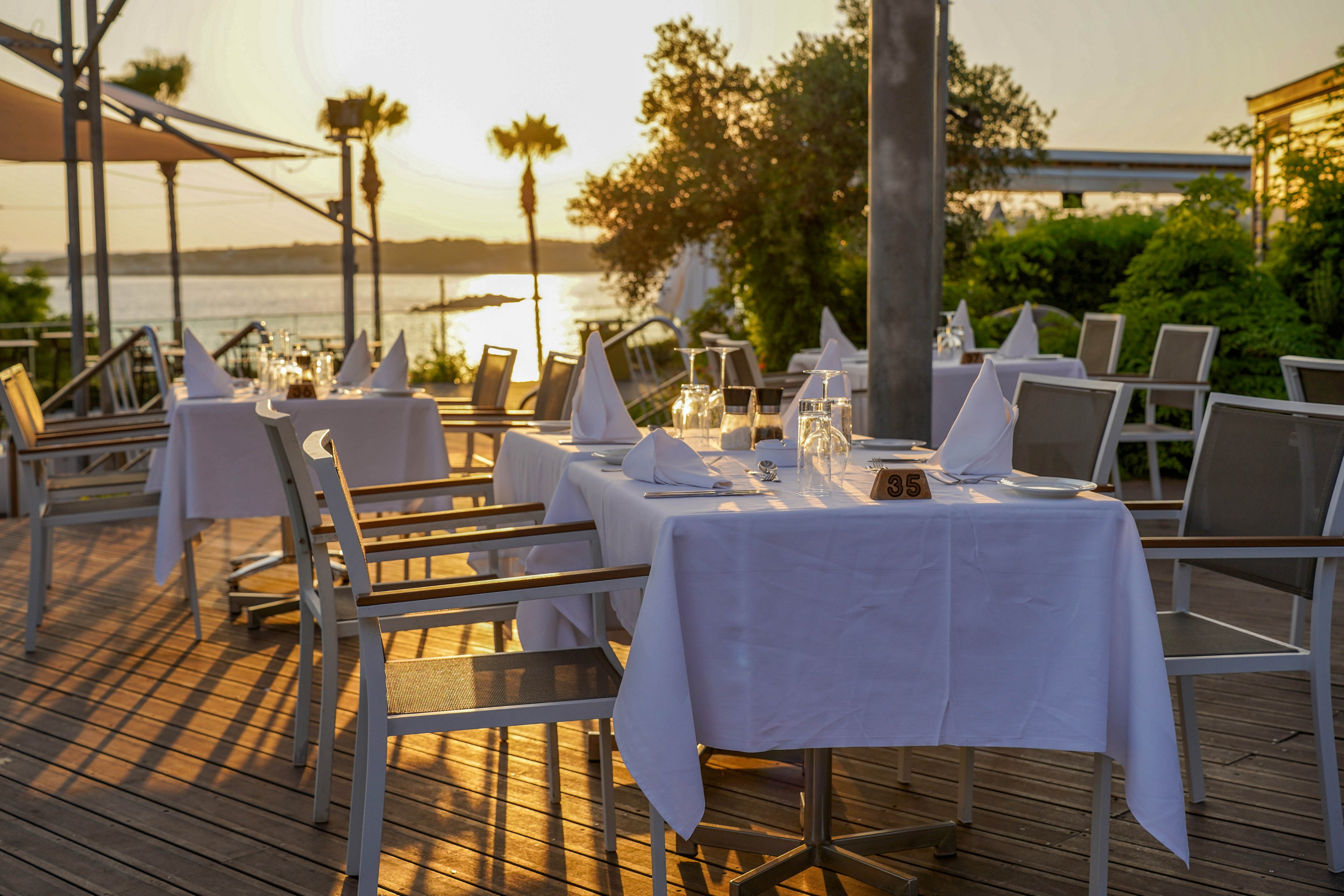 An outdoor restaurant terrace is set for dinner with white tablecloths and neatly folded napkins. Empty tables and chairs overlook a calm body of water at sunset. Warm golden light reflects off the glasses and cutlery, creating a relaxed and elegant atmosphere. Lush greenery and palm trees frame the view, enhancing the coastal, vacation-like setting.