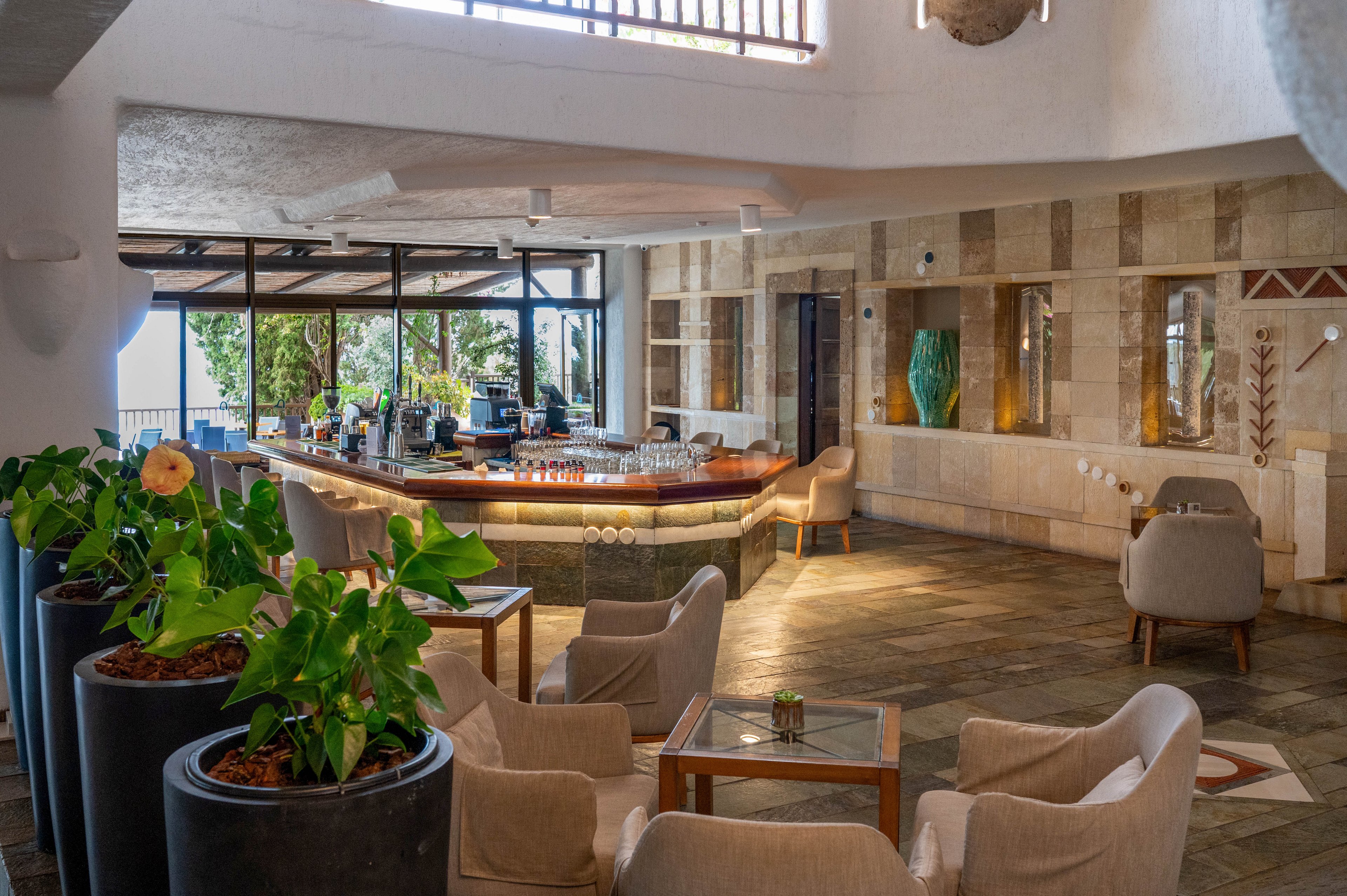 A contemporary hotel lounge bar features a curved wooden counter with neatly arranged glassware and bottles. Comfortable armchairs and small tables create an inviting seating area on a stone-tiled floor. Large windows open to views of greenery and natural light, enhancing the airy atmosphere. Decorative plants and sculptural vases add a stylish touch to the warm, neutral interior.