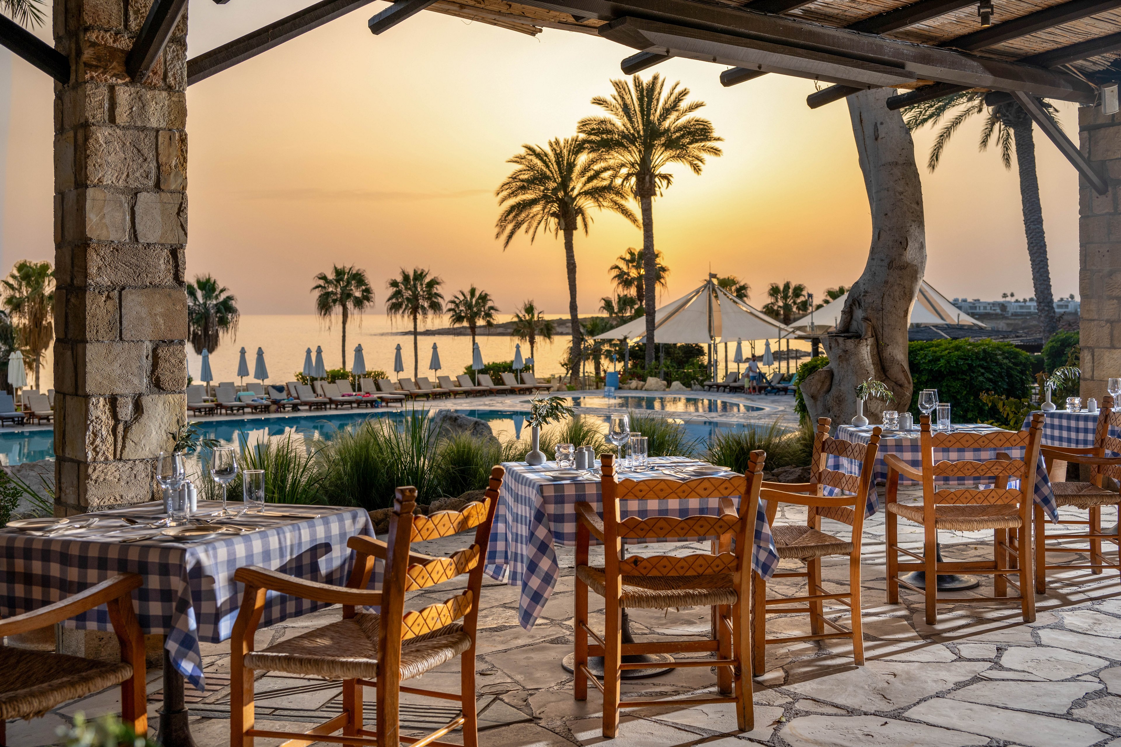 An open-air restaurant terrace overlooks a resort pool at sunset. Wooden chairs and tables with blue-and-white checkered tablecloths are set with glassware and cutlery, ready for diners. In the background, palm trees, sun loungers, and umbrellas line the pool and frame the calm sea horizon. Warm golden light creates a tranquil, romantic atmosphere ideal for evening dining by the water.