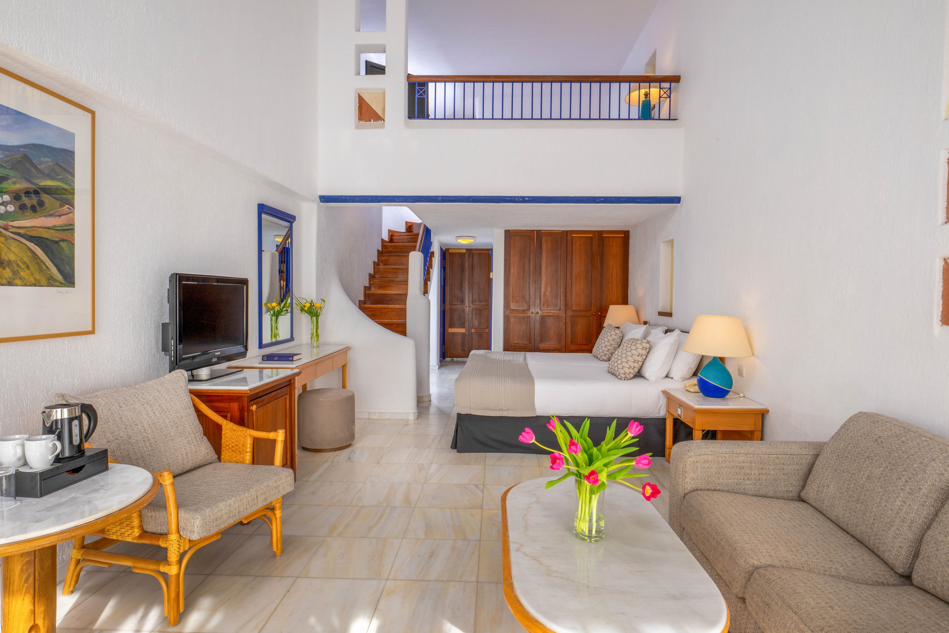 A spacious duplex hotel suite features a comfortable seating area with a sofa and coffee table in the foreground. A bed with cushions is positioned near wooden wardrobes and a staircase leading to an upper loft level. The room includes a television on a wooden console, a small dining chair, and a coffee and tea station. White walls and tiled floors enhance the airy feel, while colorful tulips and framed artwork add decorative accents. Warm table lamps and natural light create a welcoming, relaxed atmosphere.