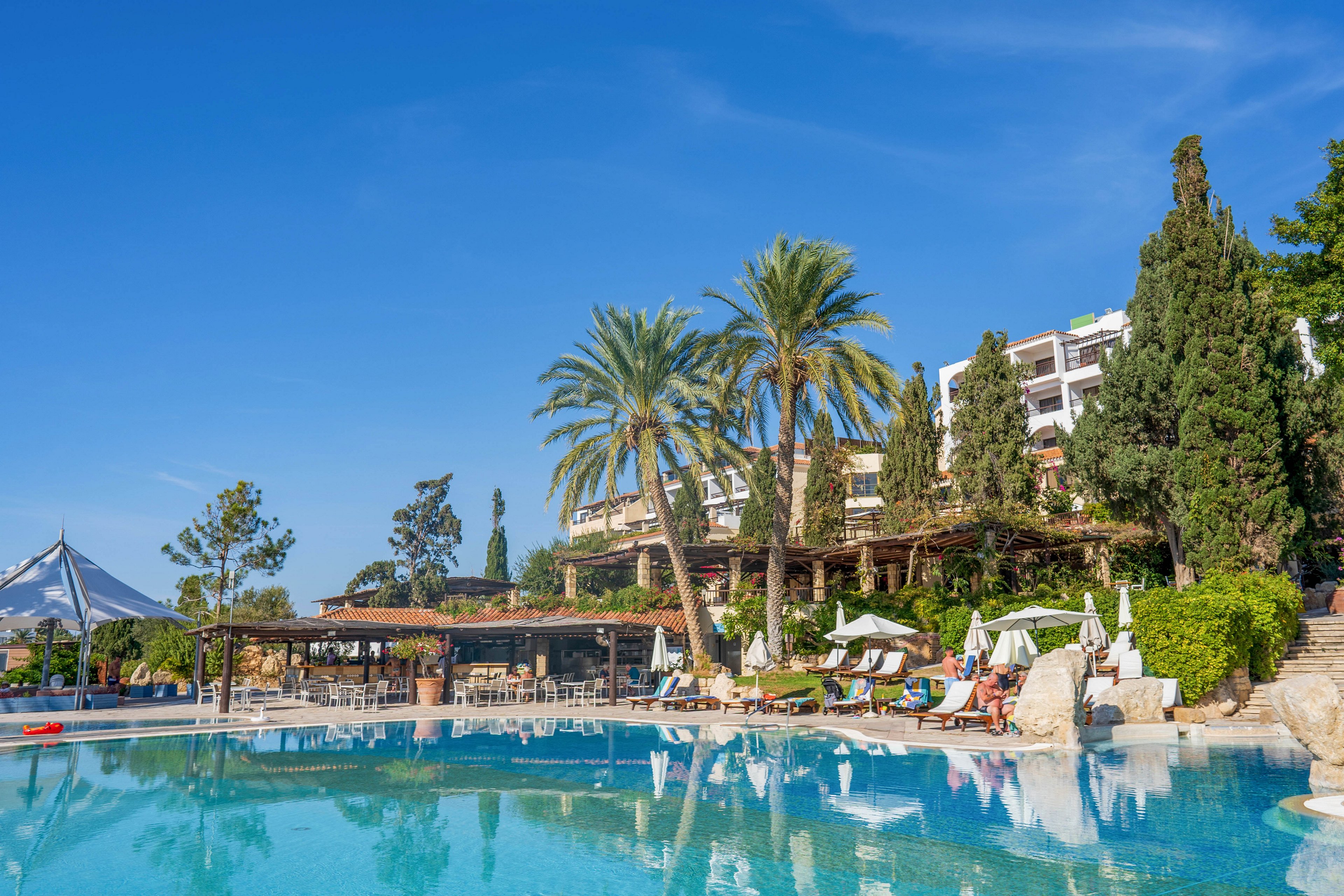 A large outdoor swimming pool reflects clear blue skies at a resort. Palm trees and lush greenery surround the pool area, creating a tropical atmosphere. Sun loungers and umbrellas line the poolside where guests relax in the shade. In the background, multi-story hotel buildings rise above the landscaped grounds, suggesting a vacation destination.