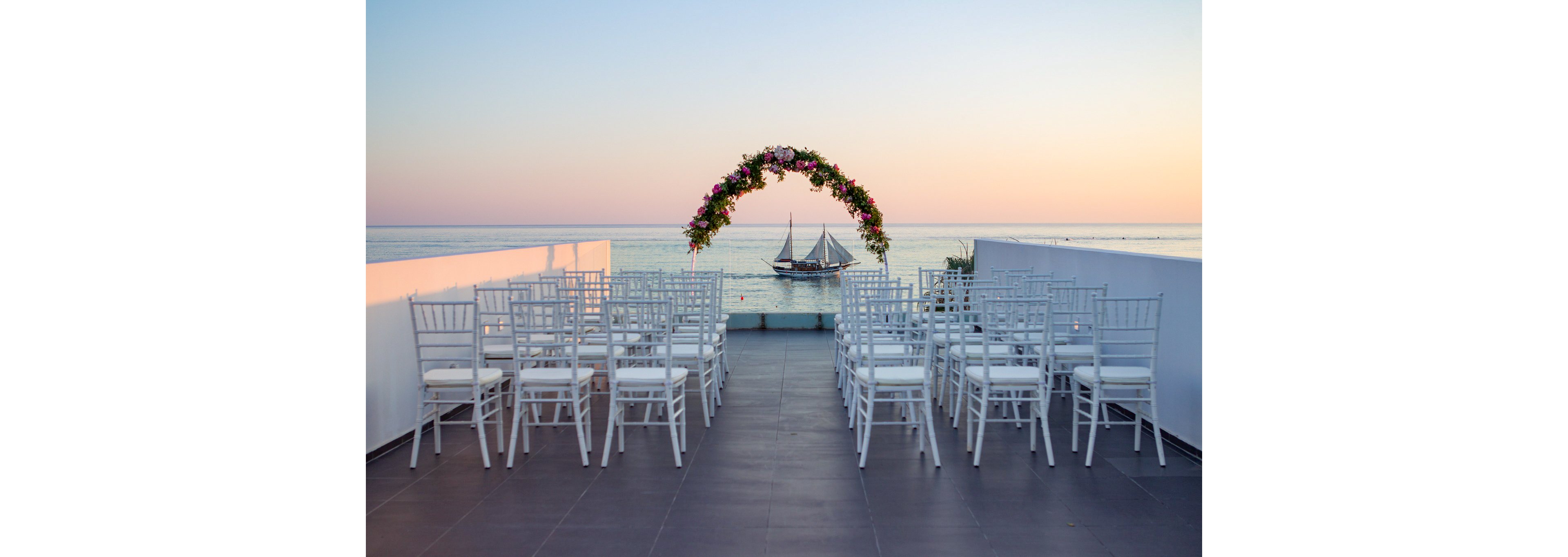 An elegant wedding ceremony setup overlooks a calm sea at sunset. Rows of white chairs face a floral arch positioned at the edge of a terrace. A sailboat glides across the water in the background beneath a pastel sky. The scene feels romantic and serene, ideal for a coastal celebration.
