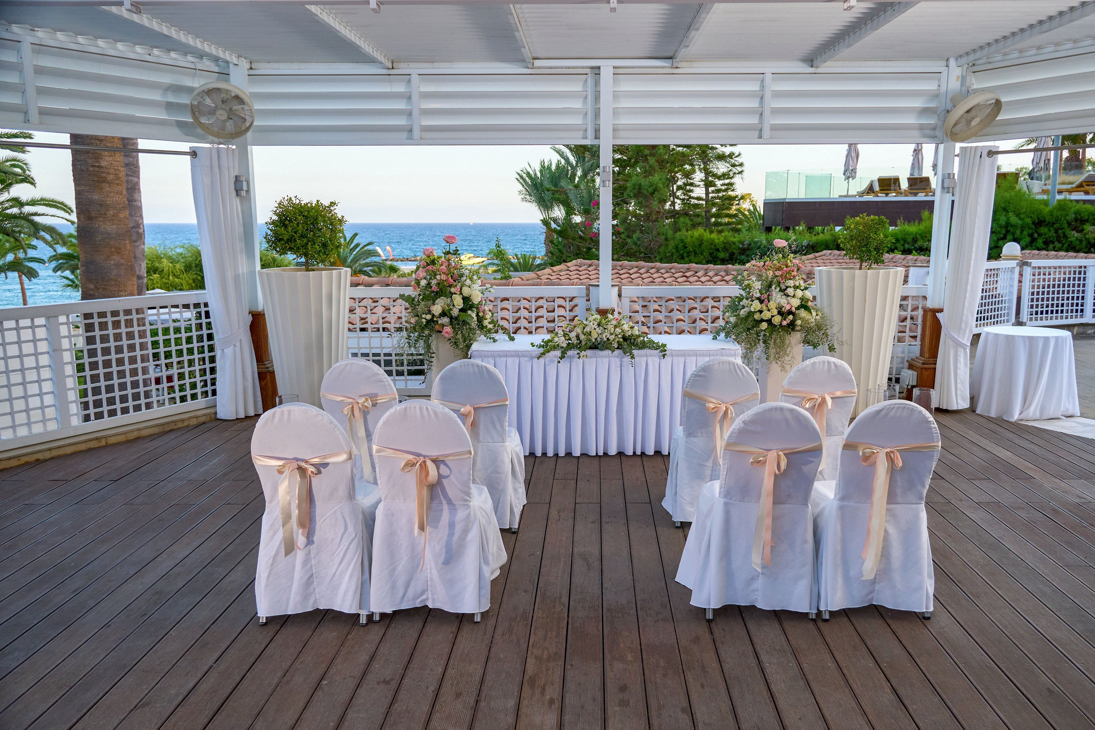 An intimate wedding ceremony setup is arranged on a wooden deck overlooking the sea. Two rows of white-covered chairs with beige ribbons face a floral-decorated altar table. Large flower arrangements and draped white curtains frame the view of the ocean and palm trees. The scene suggests a calm, romantic atmosphere suitable for a coastal wedding or vow renewal.