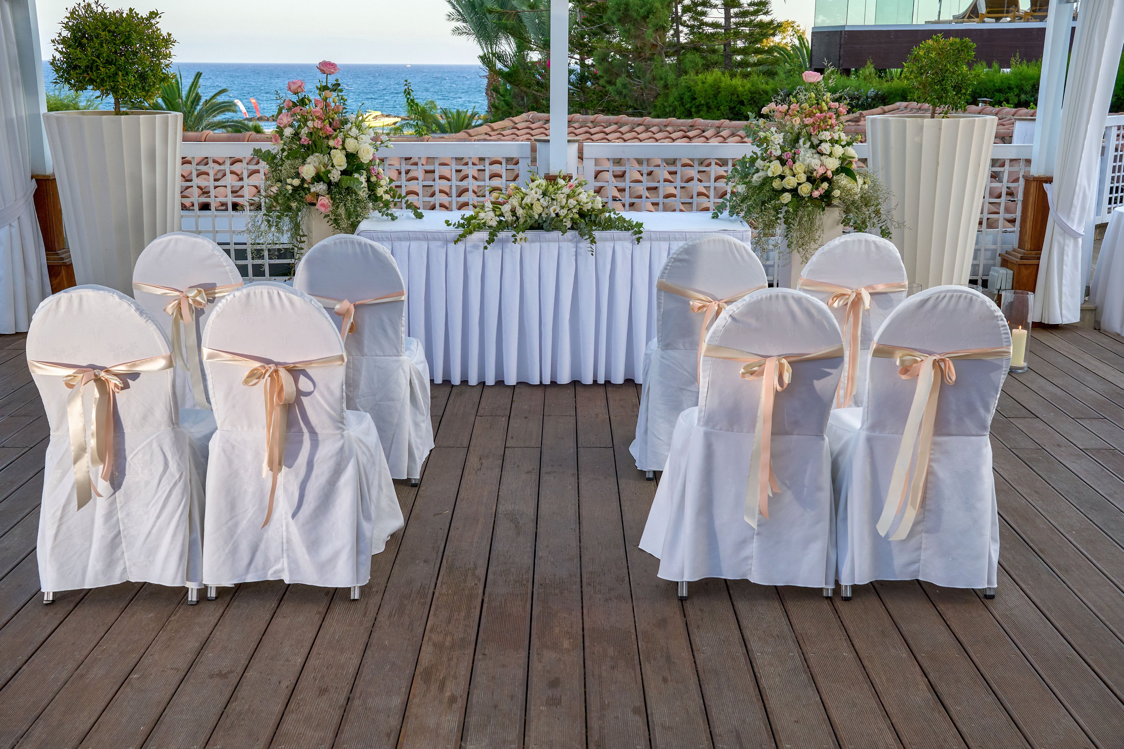An intimate wedding ceremony setup features two rows of white-covered chairs adorned with soft beige ribbons. The chairs face a long white-draped table decorated with lush floral arrangements in pastel colors. Tall flower stands and white columns frame the space, which is set on a wooden deck. In the background, the sea and coastal buildings create a serene, romantic atmosphere.