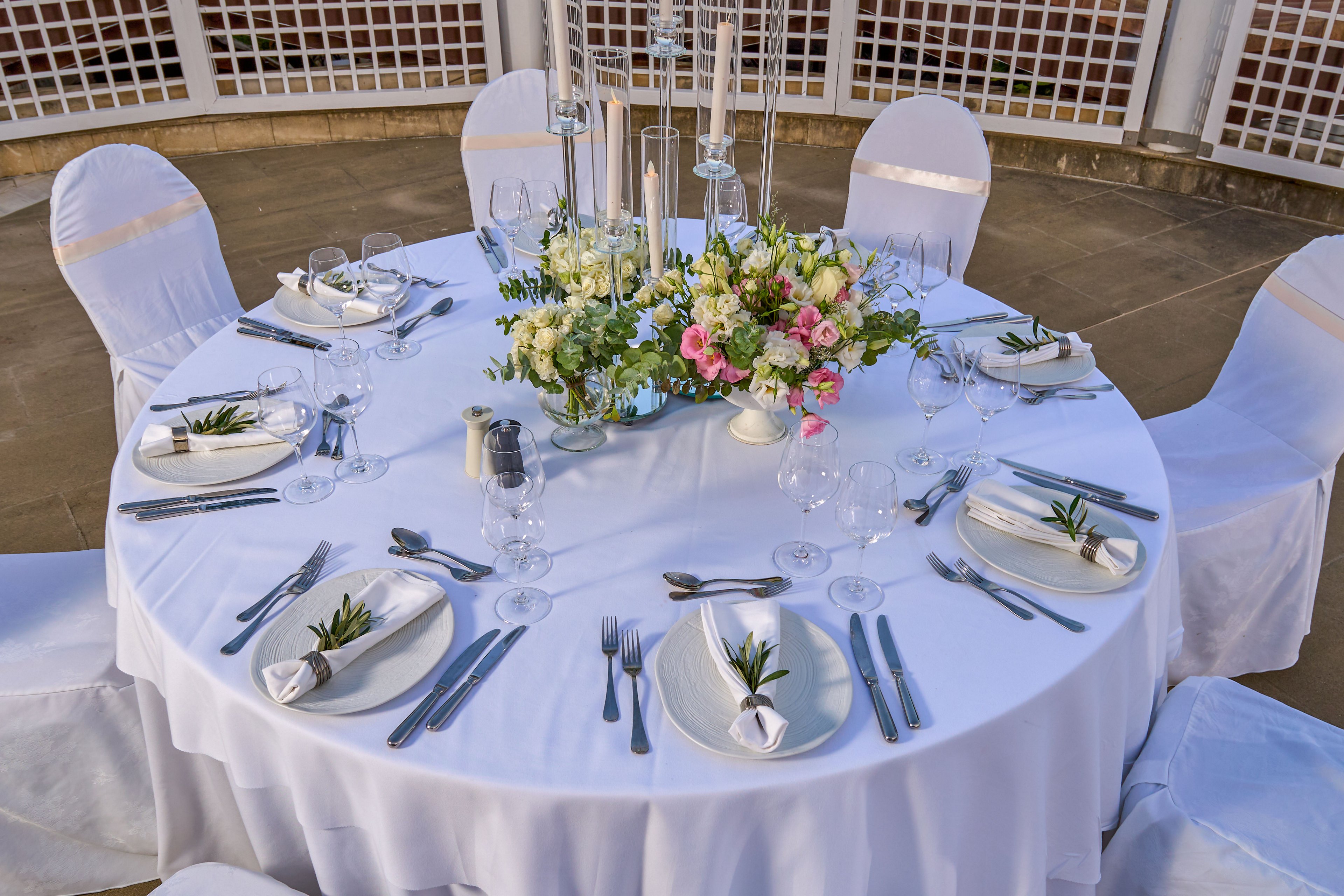 A round table is elegantly set for a formal event or wedding reception. White linens, covered chairs, and neatly arranged plates, cutlery, and glassware create a refined dining scene. A lush floral centerpiece with white and pink flowers and greenery decorates the middle of the table. Small sprigs of greenery are placed on each folded napkin, adding a natural touch to the sophisticated outdoor setting.