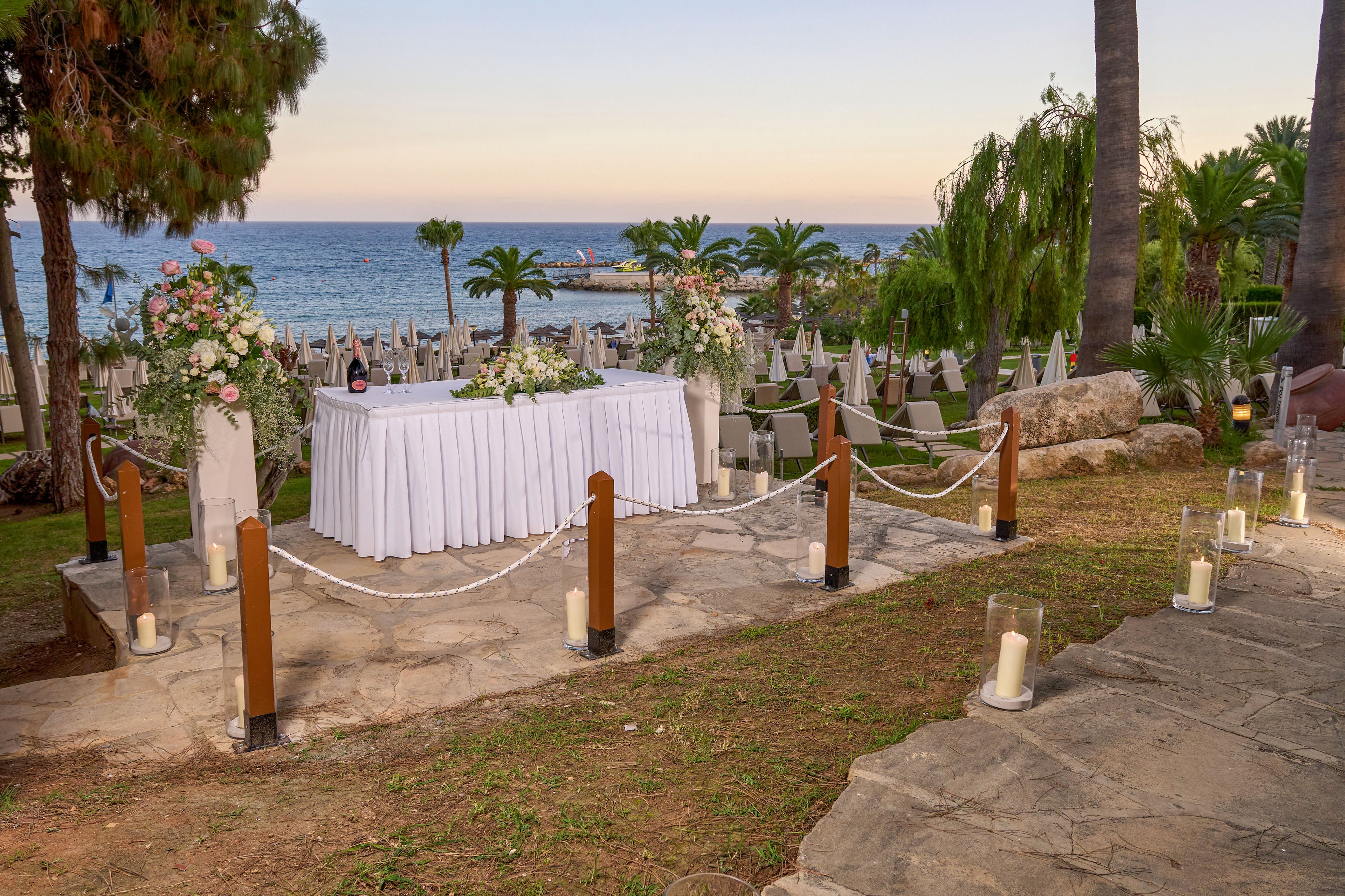 An elegant wedding reception table is arranged outdoors overlooking the sea at sunset. A long white table is decorated with lush floral arrangements and a bottle of wine or champagne. Rows of chairs and tables are set up on the terrace below, framed by palm trees and greenery. Candles in glass holders line the stone pathway, creating a romantic and intimate atmosphere. The calm ocean and pastel sky provide a serene coastal backdrop.