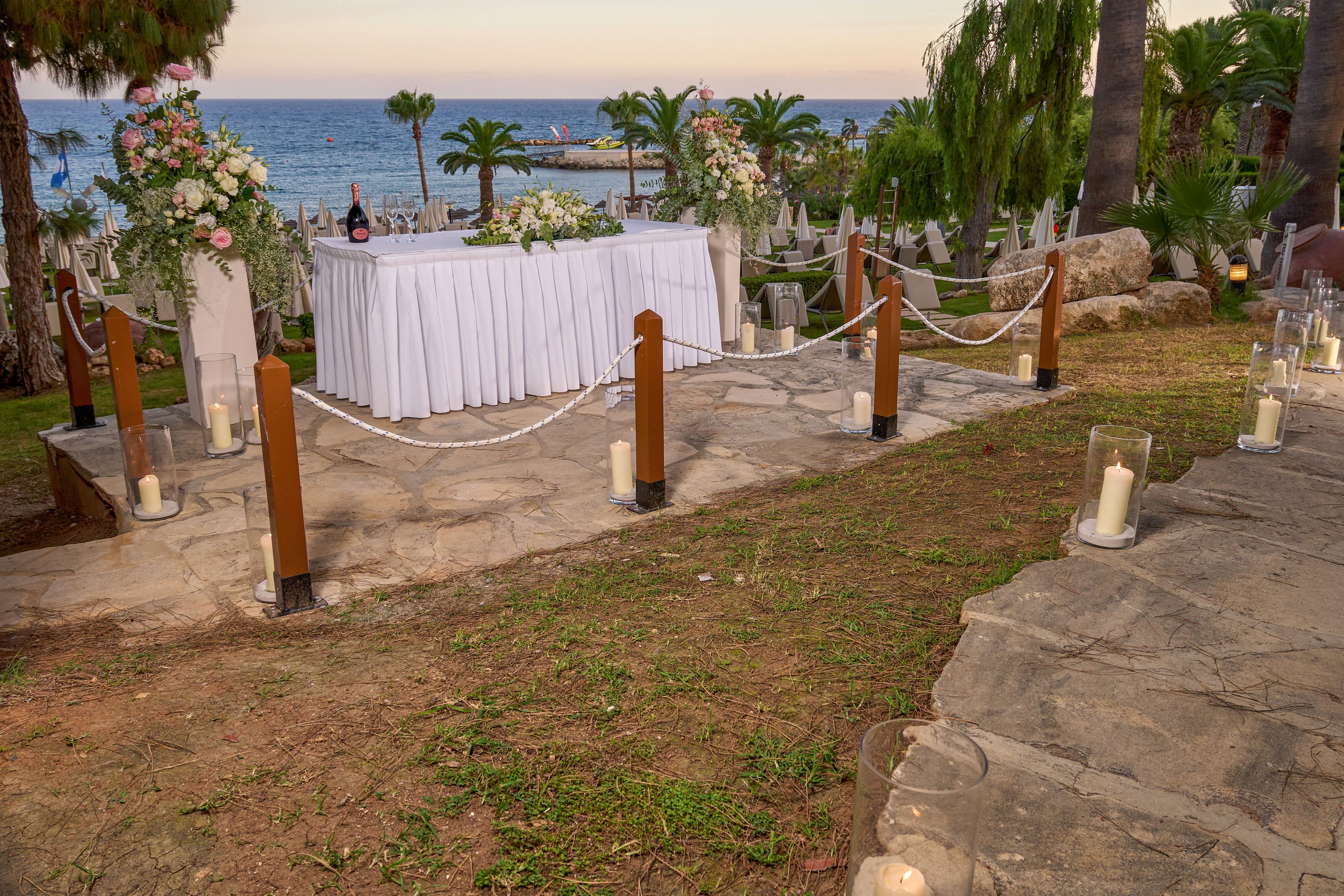 An elegant outdoor wedding ceremony setup overlooks the sea at sunset. A white-draped table decorated with lush floral arrangements and champagne bottles stands at the center. Wooden posts with white ropes frame the stone pathway, while glass candle lanterns line the steps and ground. Palm trees, greenery, and calm ocean water create a romantic coastal backdrop.