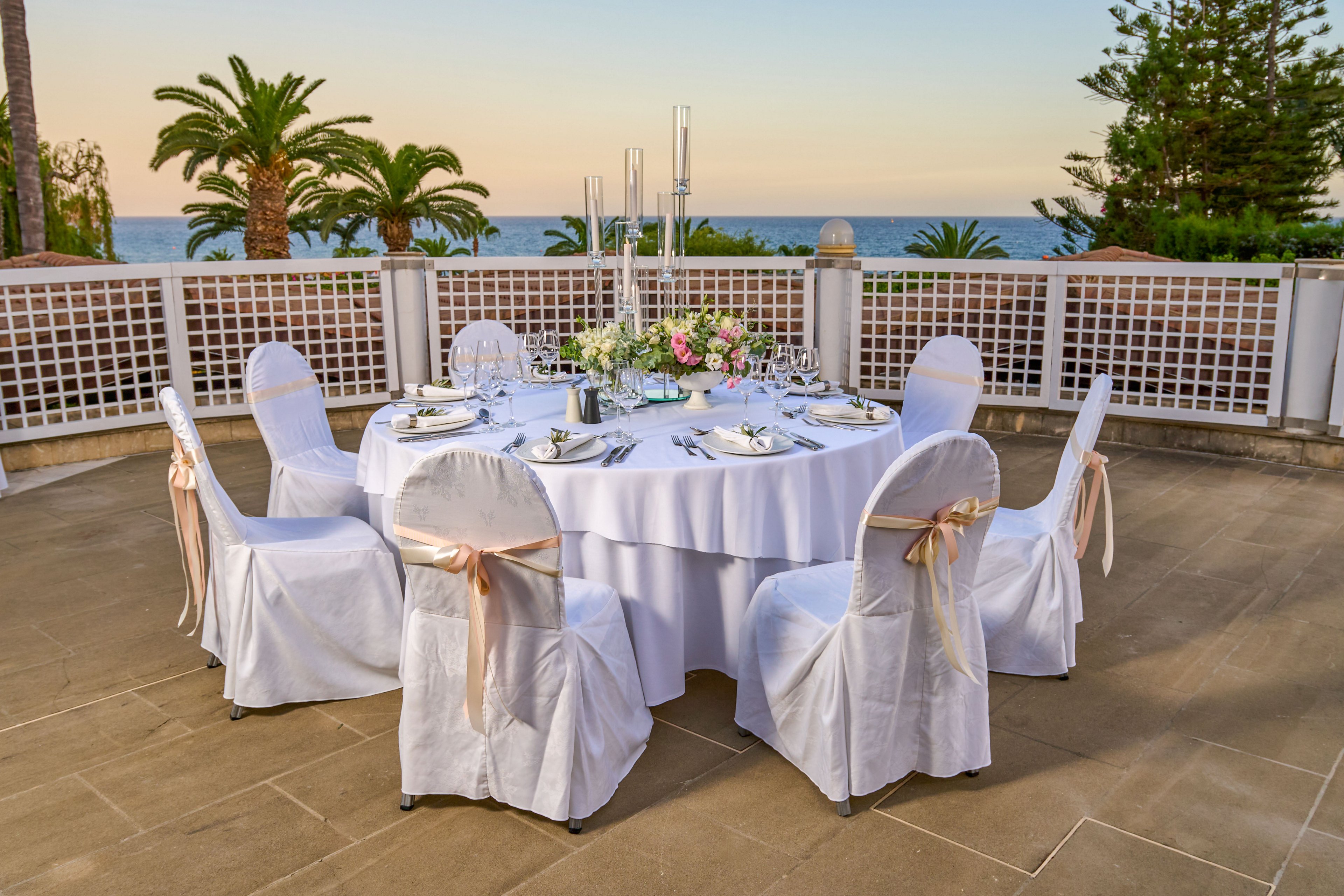 A round table is elegantly set for a formal event on a terrace overlooking the sea. White linens, covered chairs with soft beige ribbons, and polished tableware create a refined atmosphere. A floral centerpiece and tall candlesticks decorate the middle of the table. Palm trees, a low fence, and a calm ocean horizon frame the scene at sunset.