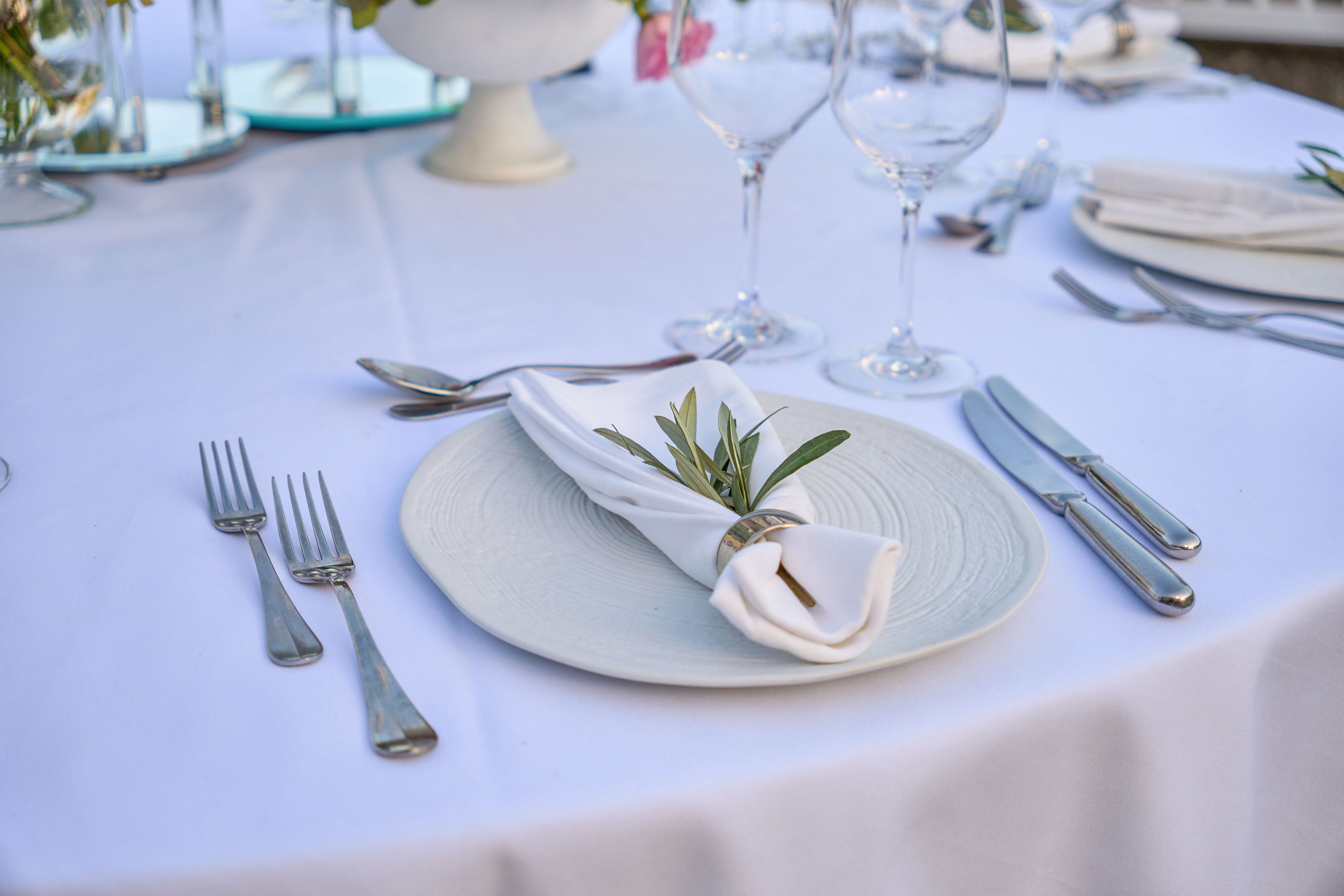A neatly arranged place setting features a white plate with a folded white napkin tied with a small sprig of greenery. Silver cutlery is carefully positioned on either side of the plate atop a crisp white tablecloth. In the background, wine glasses and decorative elements suggest a formal dining occasion. The overall scene conveys a refined, minimalist style suitable for weddings or upscale events.
