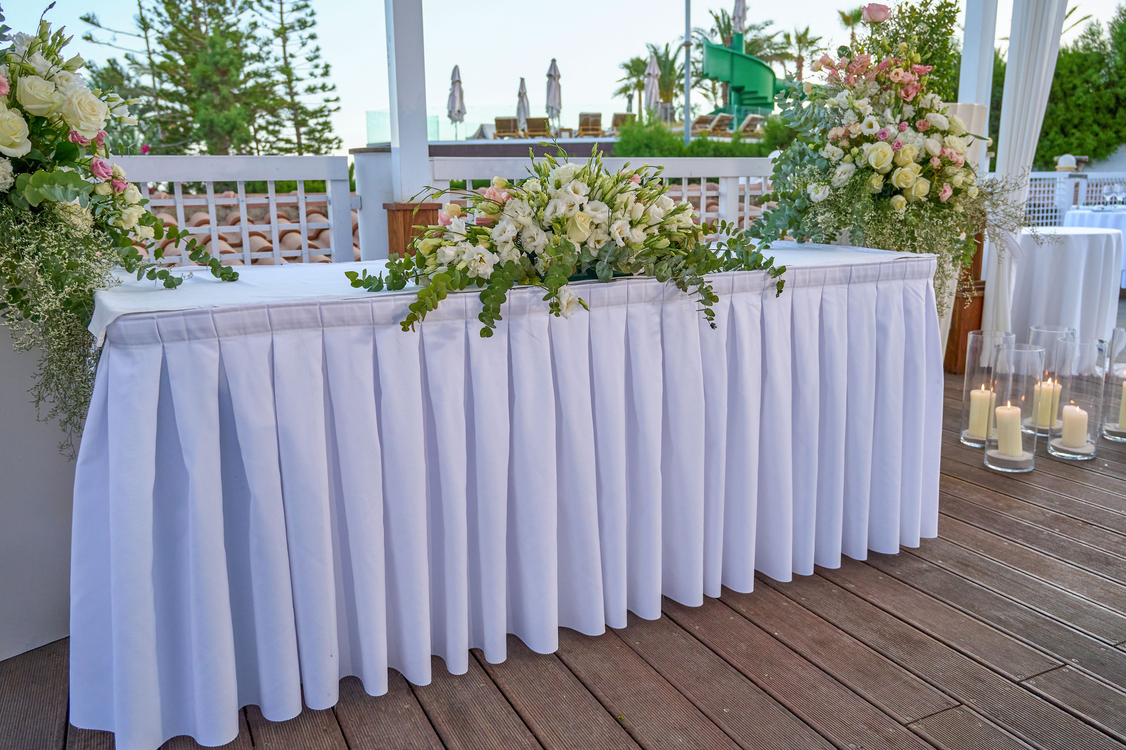 A long white table is beautifully decorated with lush floral arrangements and greenery. The table is set on a wooden deck in an outdoor venue, framed by white railings and distant trees. Soft candlelight from tall glass hurricanes adds a romantic glow to the scene. The overall setting suggests a sophisticated wedding or special event in a serene, coastal-like environment.