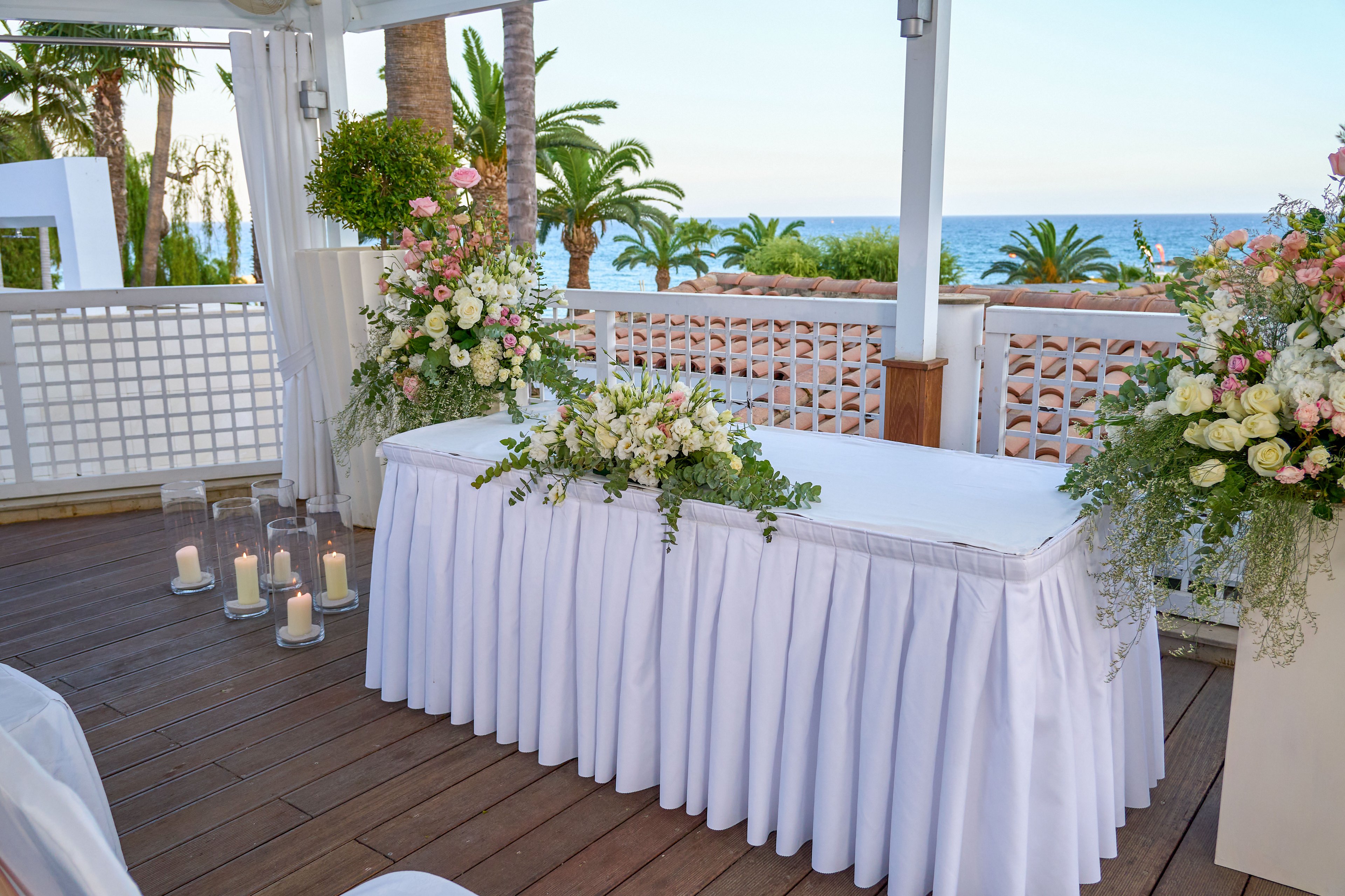 A white-draped table decorated with lush floral arrangements is set up on a wooden deck overlooking the sea. Tall glass candle holders with lit candles stand beside the table, adding a romantic glow. The setting features white railings, greenery, and palm trees framing the ocean view. Soft natural light and pastel flowers create a serene and elegant wedding atmosphere.