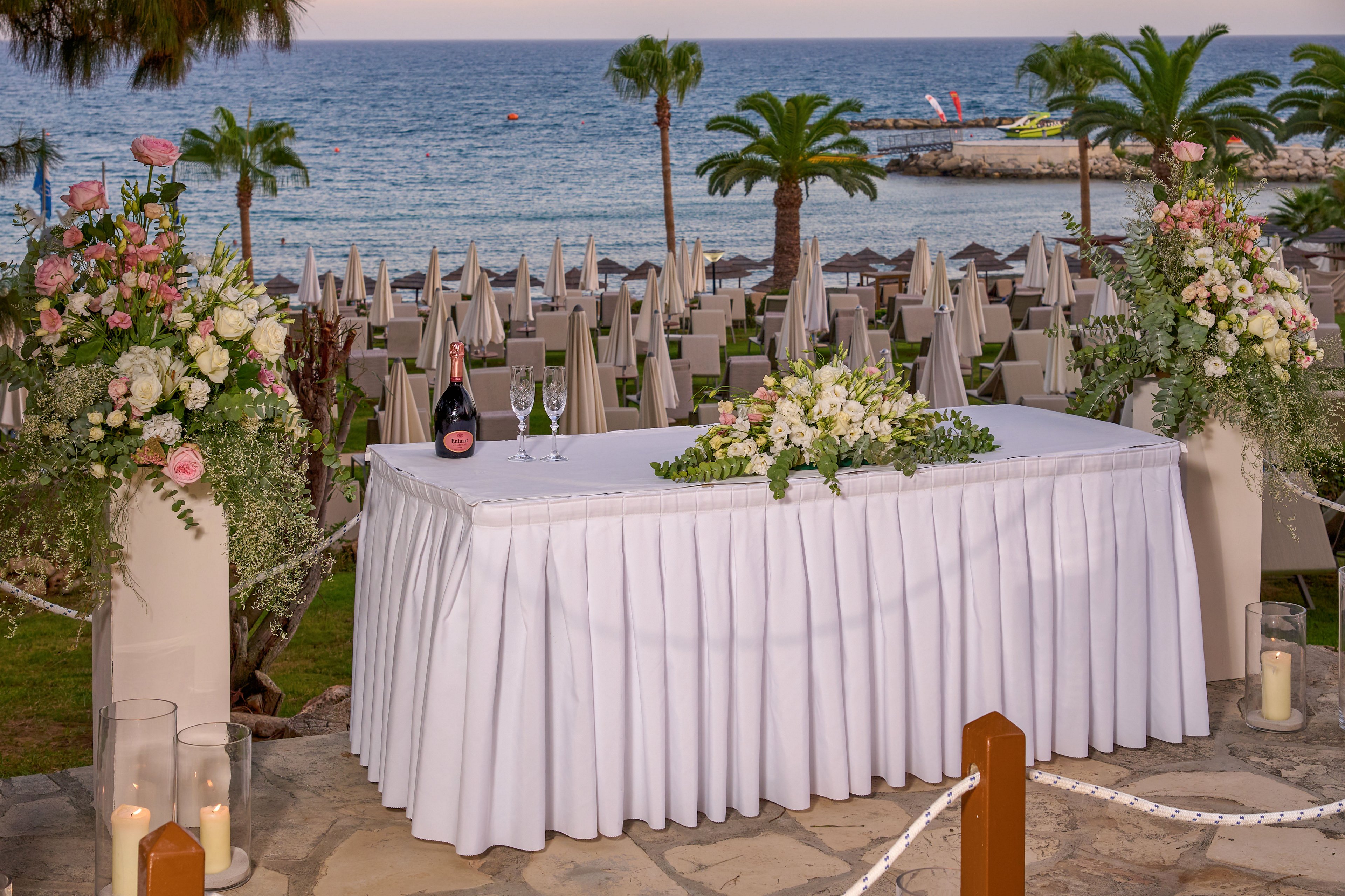 A long white-draped table is beautifully arranged for a seaside wedding reception. The table is decorated with lush floral arrangements, greenery, and a bottle of sparkling wine with two glasses. Tall flower stands and candles in glass holders frame the scene, adding a romantic touch. In the background, rows of chairs and umbrellas line the beachfront, with palm trees and calm sea water completing the picturesque coastal setting.