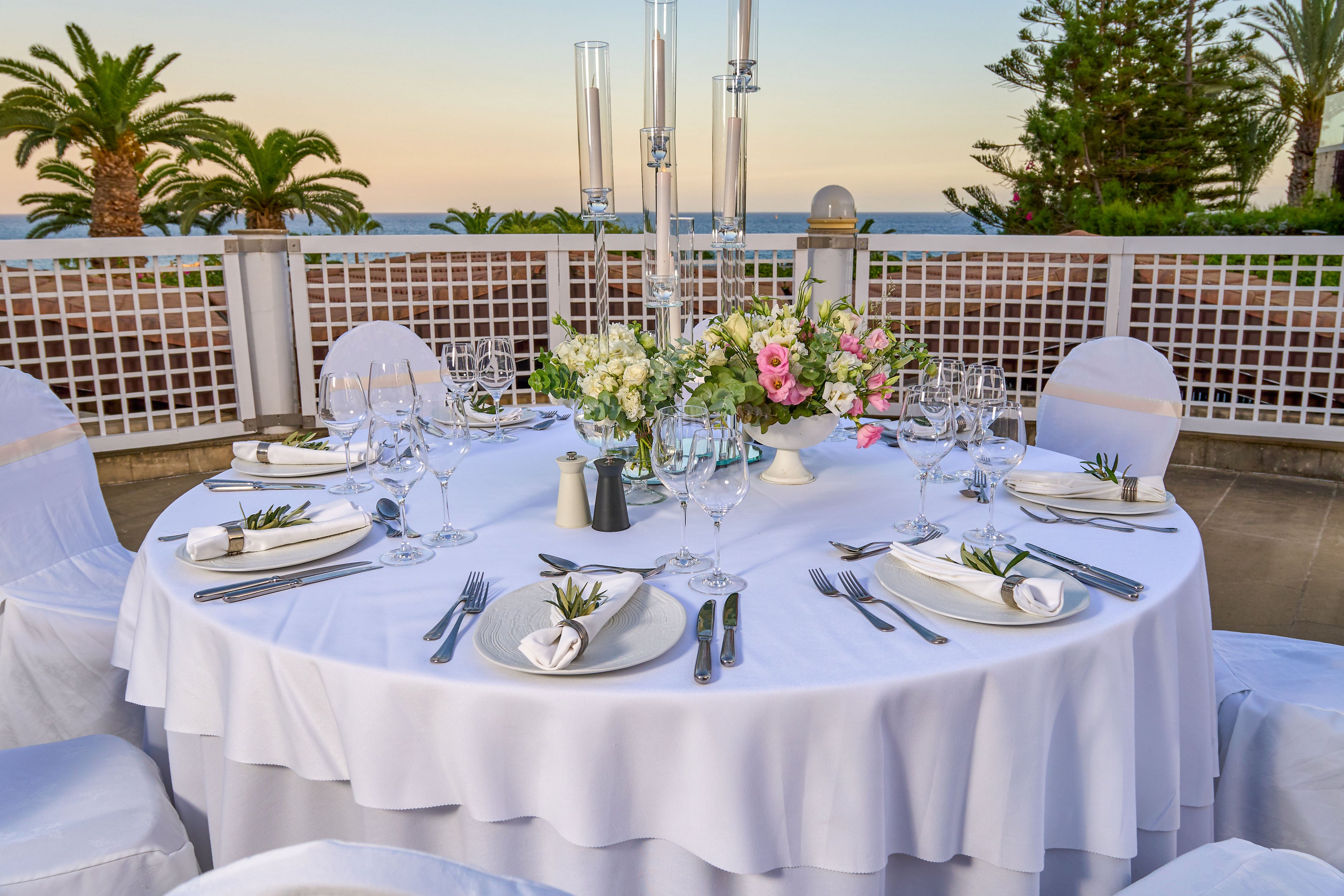 A round table is elegantly set for a formal outdoor event, featuring white linens and neatly folded napkins. Multiple wine glasses, plates, and cutlery are arranged precisely around each place setting. A floral centerpiece with white and pink flowers decorates the middle of the table, adding a romantic touch. The scene overlooks a terrace with palm trees and a distant ocean view at sunset, creating a serene and festive atmosphere.