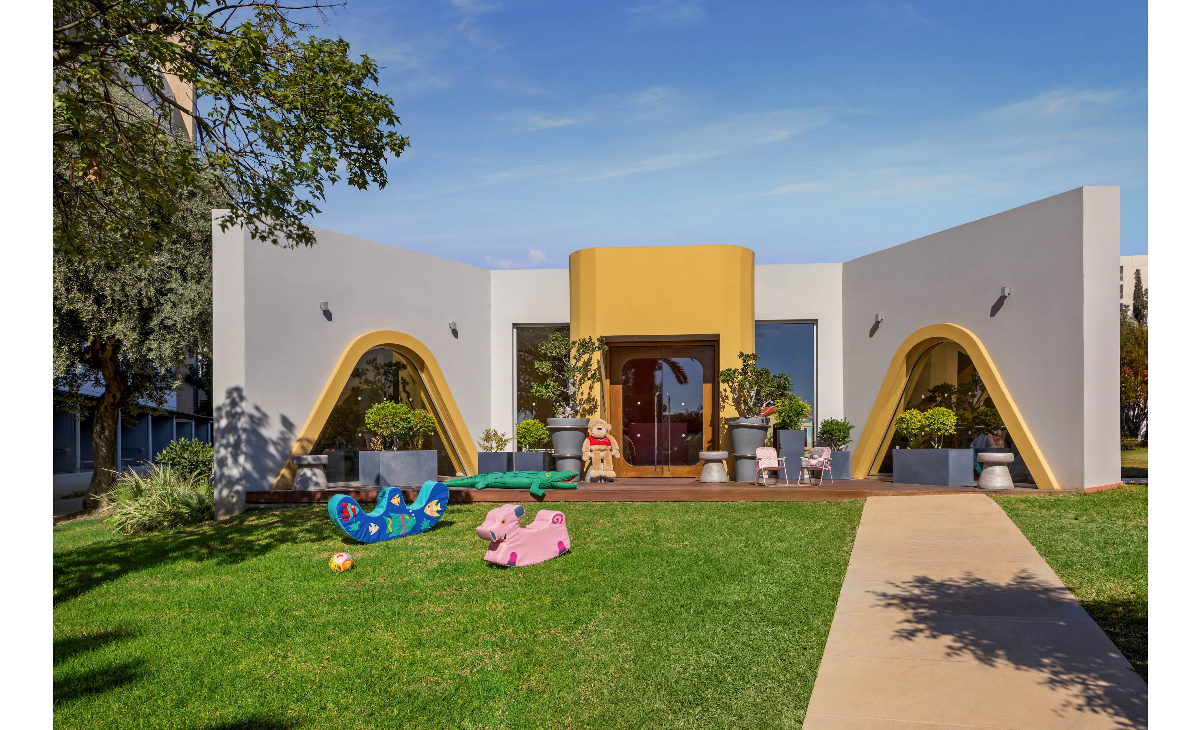 A modern, single-story building with bold geometric arches and a yellow entrance stands behind a neatly manicured lawn. The setting appears to be a daycare or preschool, with several colorful rocking toys and a small slide scattered across the grass. Large potted plants frame the doorway, and expansive windows reflect the clear blue sky. A concrete path leads from the foreground toward the entrance, creating a welcoming and family-friendly atmosphere.
