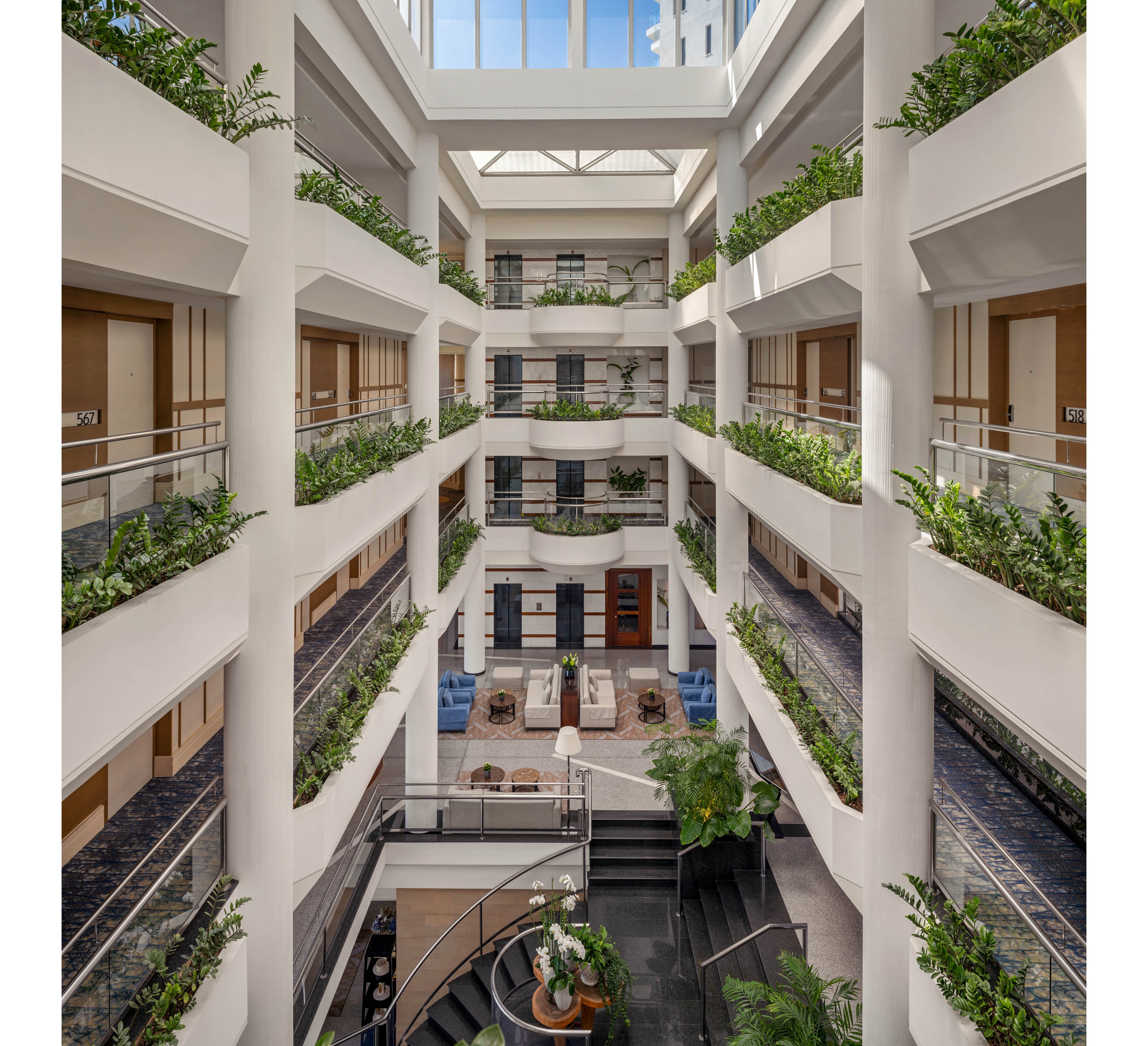 A multi-story hotel atrium features open interior corridors overlooking a central lounge area. Each floor has railings lined with lush green plants, creating a vertical garden effect. Natural light streams in from large skylights above, illuminating the white structural columns and walkways. The ground level includes seating areas and decorative plants, giving the space a bright and welcoming atmosphere.