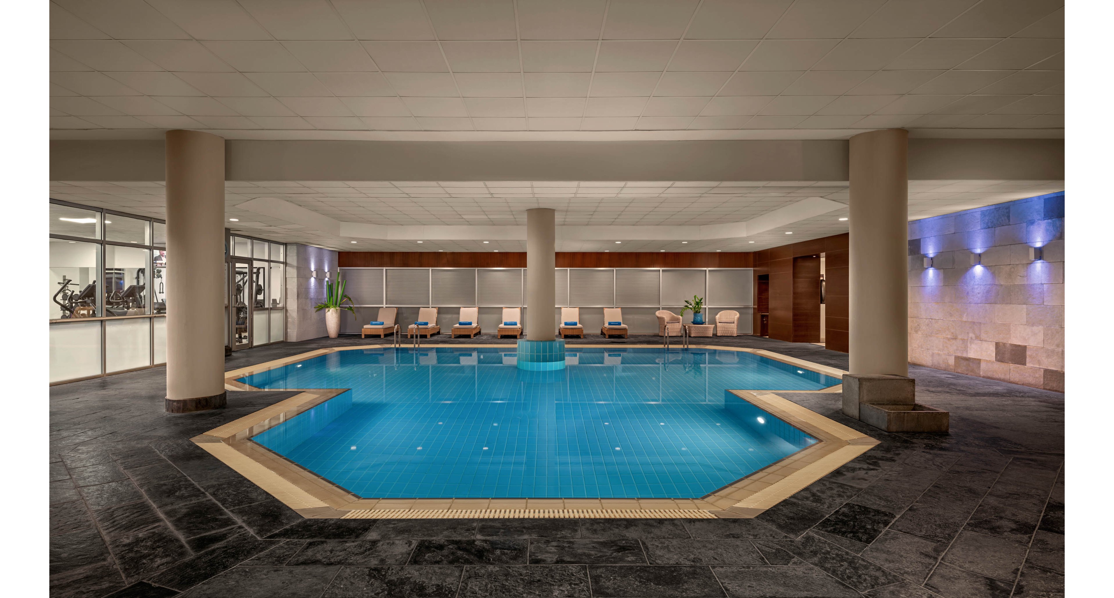 A spacious indoor swimming pool is centered in a contemporary hotel or spa setting. The octagonal pool is surrounded by dark stone flooring and neutral-toned walls, creating a clean and modern look. Several white lounge chairs line the far side of the pool, with potted plants adding a touch of greenery. Soft ceiling and wall lighting reflects on the water, enhancing the calm and relaxing atmosphere.