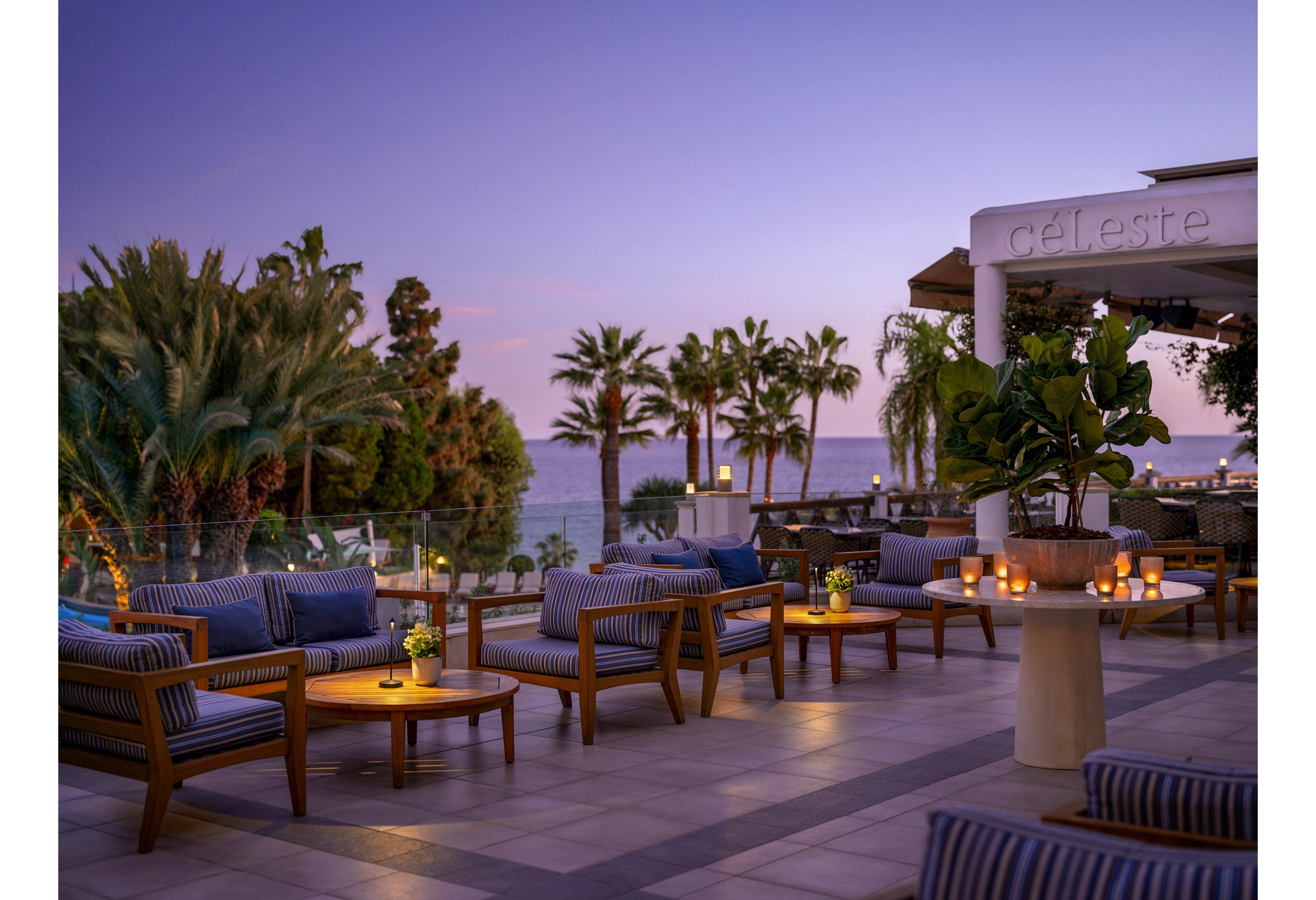 An elegant outdoor lounge terrace overlooks the sea at dusk, with comfortable striped sofas and low wooden tables. Soft candlelight glows on the tables, creating a warm and inviting atmosphere against the purple and pink evening sky. Palm trees and lush greenery frame the view, while a modern bar or restaurant area is visible in the background. The scene suggests a relaxed coastal resort setting ideal for evening drinks or social gatherings.