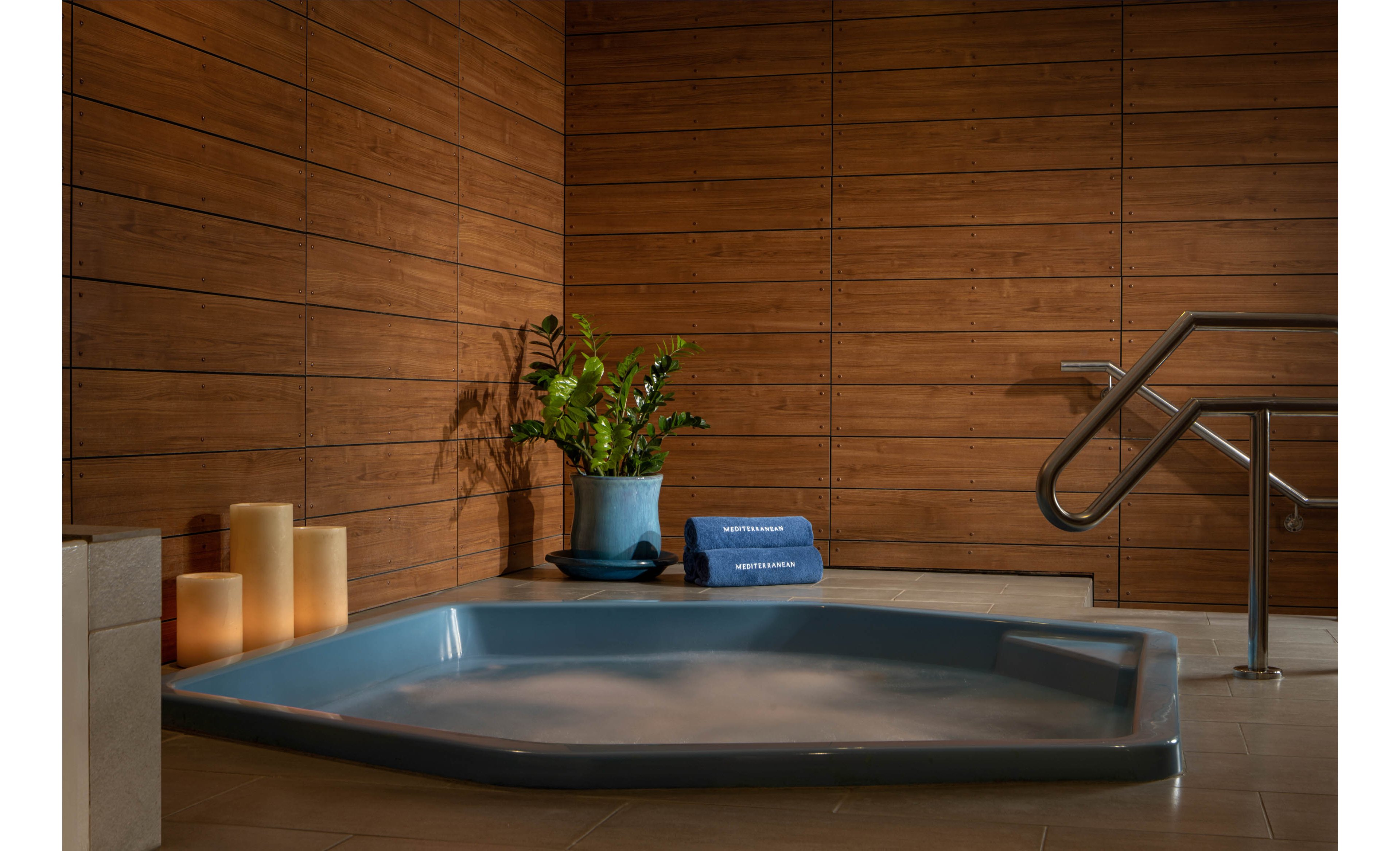 A modern indoor spa features a blue, recessed hot tub filled with gently steaming water. The tub is set against a warm wooden paneled wall, creating a cozy and contemporary atmosphere. A potted green plant and rolled blue towels rest on the edge of the tub, adding a touch of color and comfort. Softly glowing pillar candles enhance the relaxing mood, while a metal handrail suggests safe and easy access to the spa.
