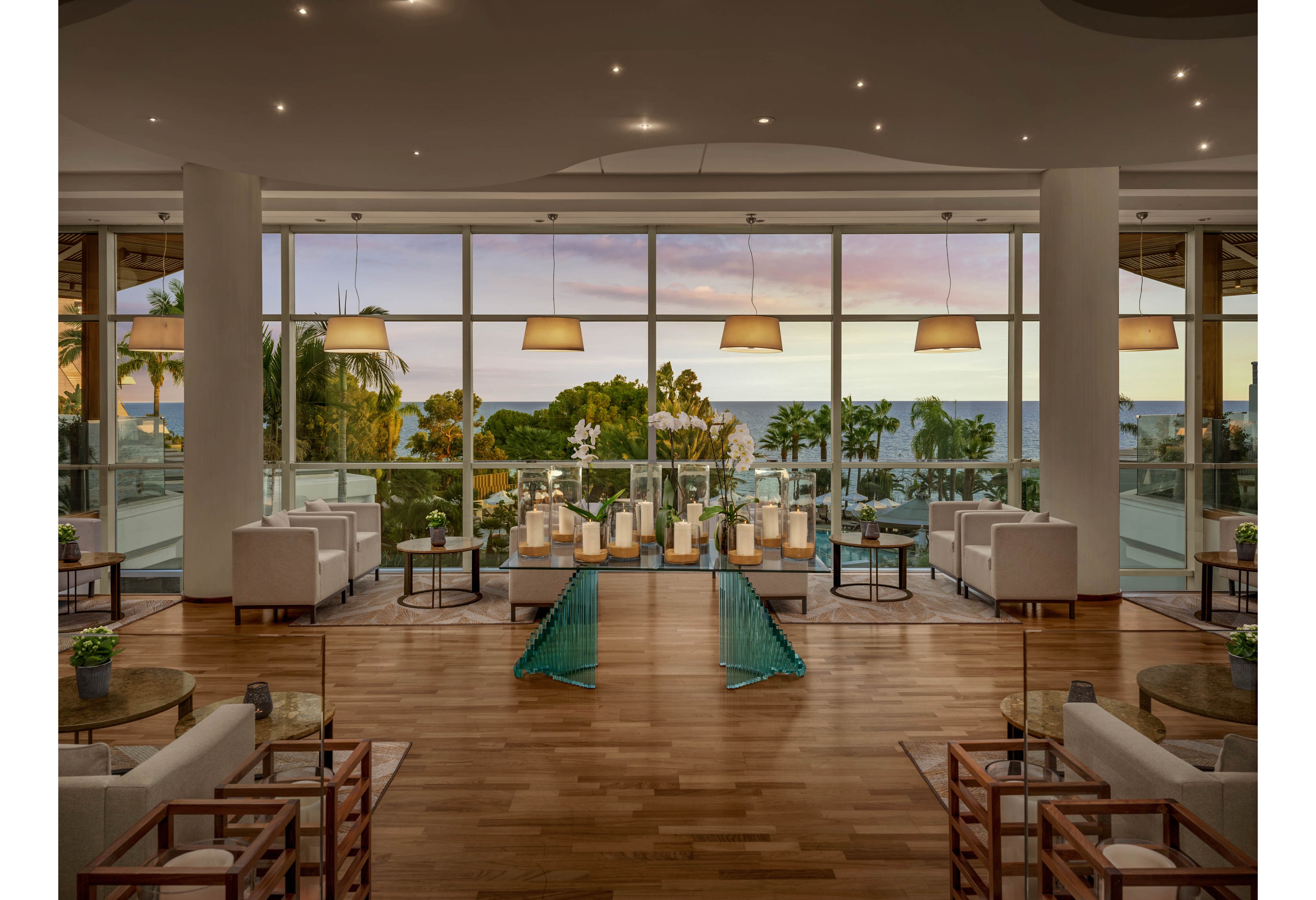 A spacious modern hotel lounge features white sofas and armchairs arranged symmetrically on a polished wooden floor. Large floor-to-ceiling windows reveal palm trees, a terrace, and an ocean view at sunset. Pendant lights and recessed ceiling lights create a warm, ambient glow throughout the room. Decorative lanterns and small potted plants add a relaxed, upscale resort atmosphere.