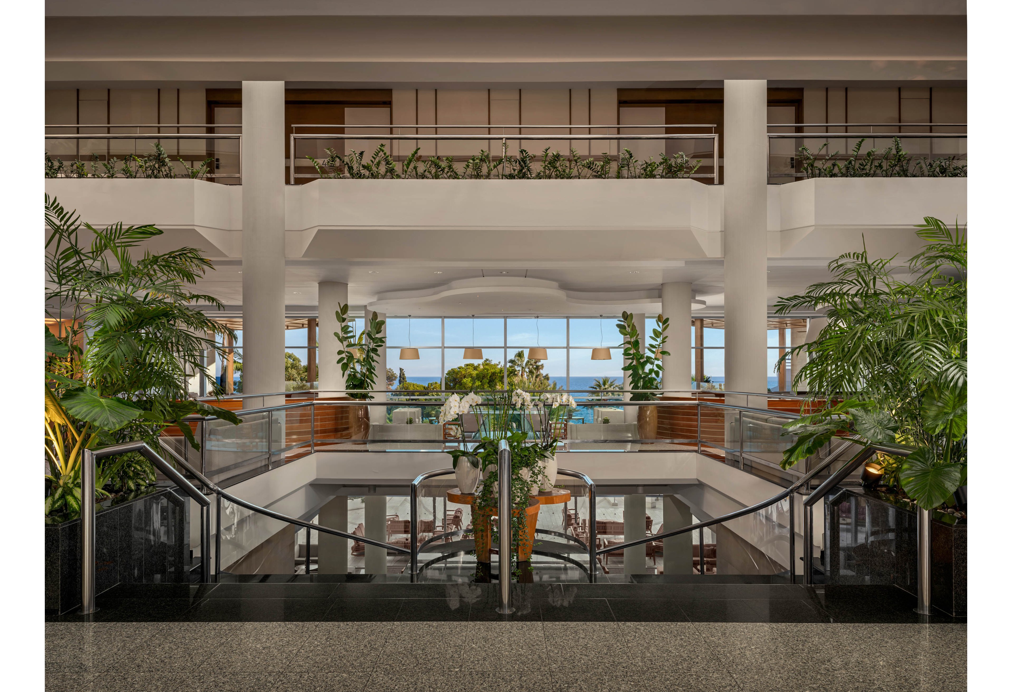 A spacious, contemporary hotel lobby features a central staircase framed by glass railings and lush green plants. Large windows at the far end reveal a bright ocean view and blue sky. Neutral tones, polished floors, and clean architectural lines create a calm, upscale atmosphere. Seating and tables are arranged on the lower level, suggesting a relaxed social or lounge area.