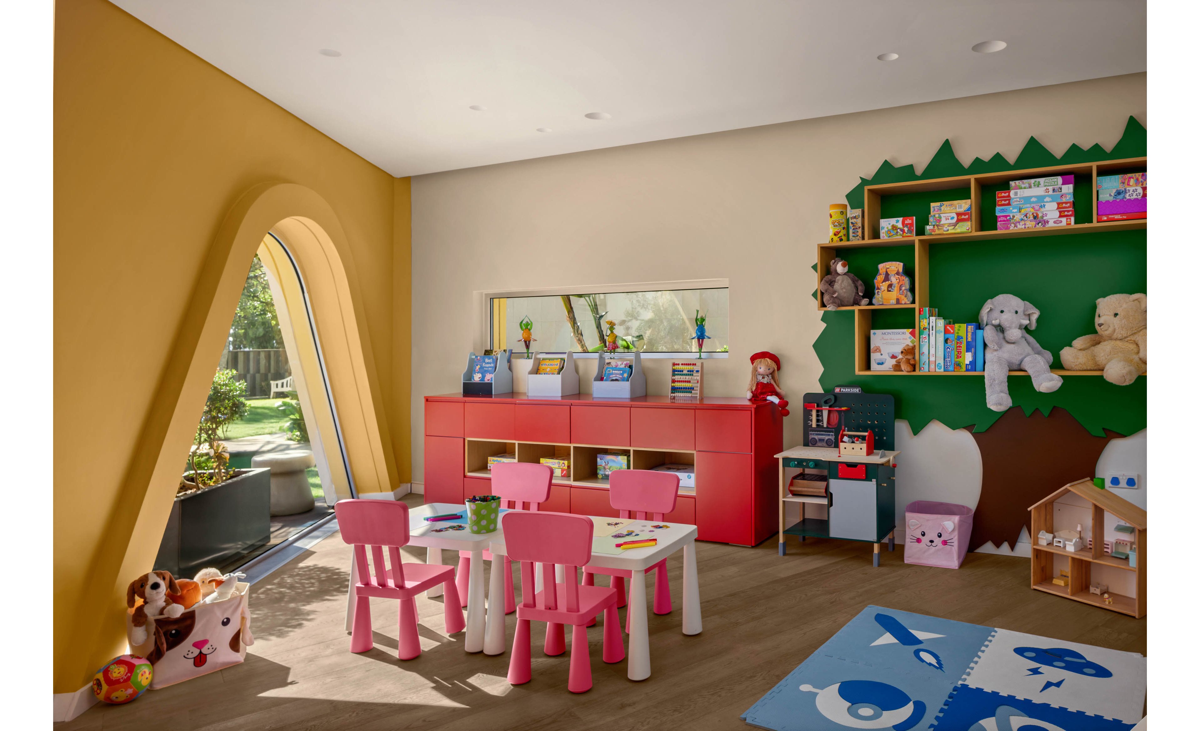 A cheerful children’s playroom features small pink chairs around white tables in the center of the room. The space includes red storage cabinets, open shelves filled with toys, books, and stuffed animals, and a dollhouse in the corner. Large windows and a distinctive arched opening bring in natural light and views of the garden outside. Soft play mats on the floor and art supplies on the tables create a welcoming environment for kids’ activities.