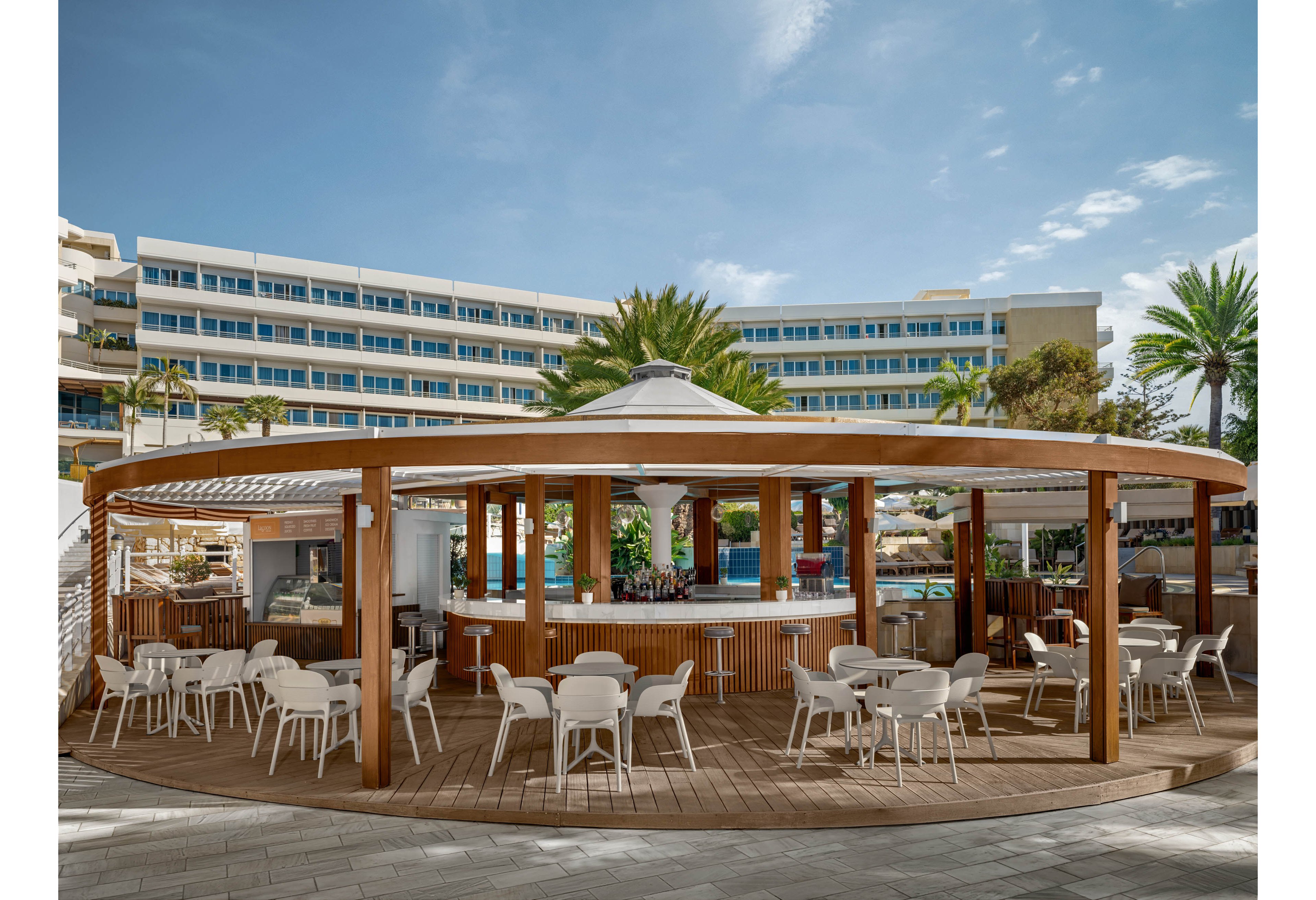 An open-air circular bar with wooden beams and a white roof stands in the foreground of a resort setting. White plastic chairs and tables surround the bar on a wooden deck, creating a casual outdoor seating area. In the background, a multi-story hotel building with rows of blue and white balconies faces the bar. Palm trees and a clear blue sky enhance the relaxed, tropical atmosphere. The scene suggests a vacation destination focused on leisure and poolside refreshments.