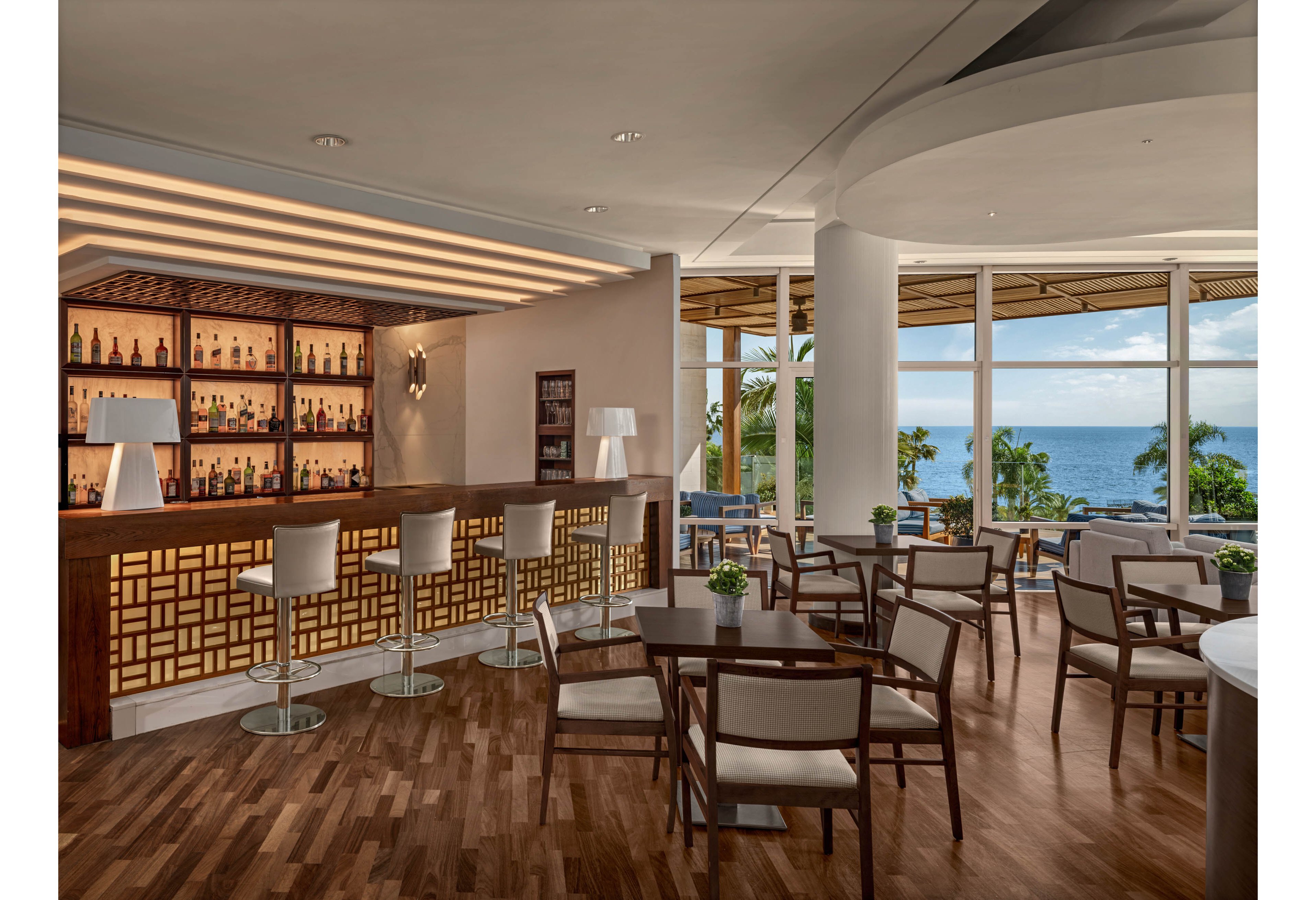 A contemporary hotel lounge bar features a wooden counter with illuminated geometric panels and a backlit wall of bottles. Sleek barstools and matching dining chairs surround small tables arranged on polished wood flooring. Large floor-to-ceiling windows reveal palm trees and a bright ocean view outside. Neutral tones, clean lines, and soft lighting create a calm, upscale atmosphere suitable for relaxing or socializing.