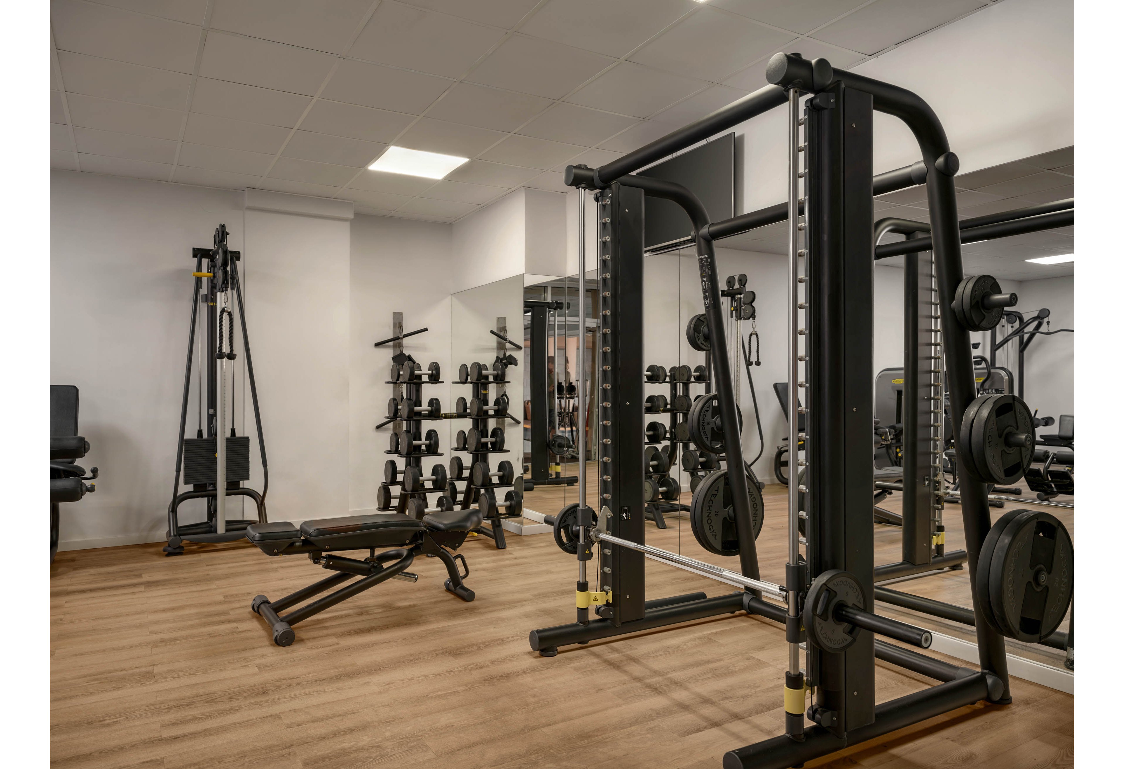 A modern fitness room features various strength training machines and free weights. A large cable machine and a squat rack dominate the foreground, while dumbbells and benches line the mirrored back wall. The space has light-colored walls, a wooden floor, and bright overhead lighting. The gym appears clean, organized, and ready for individual workouts.