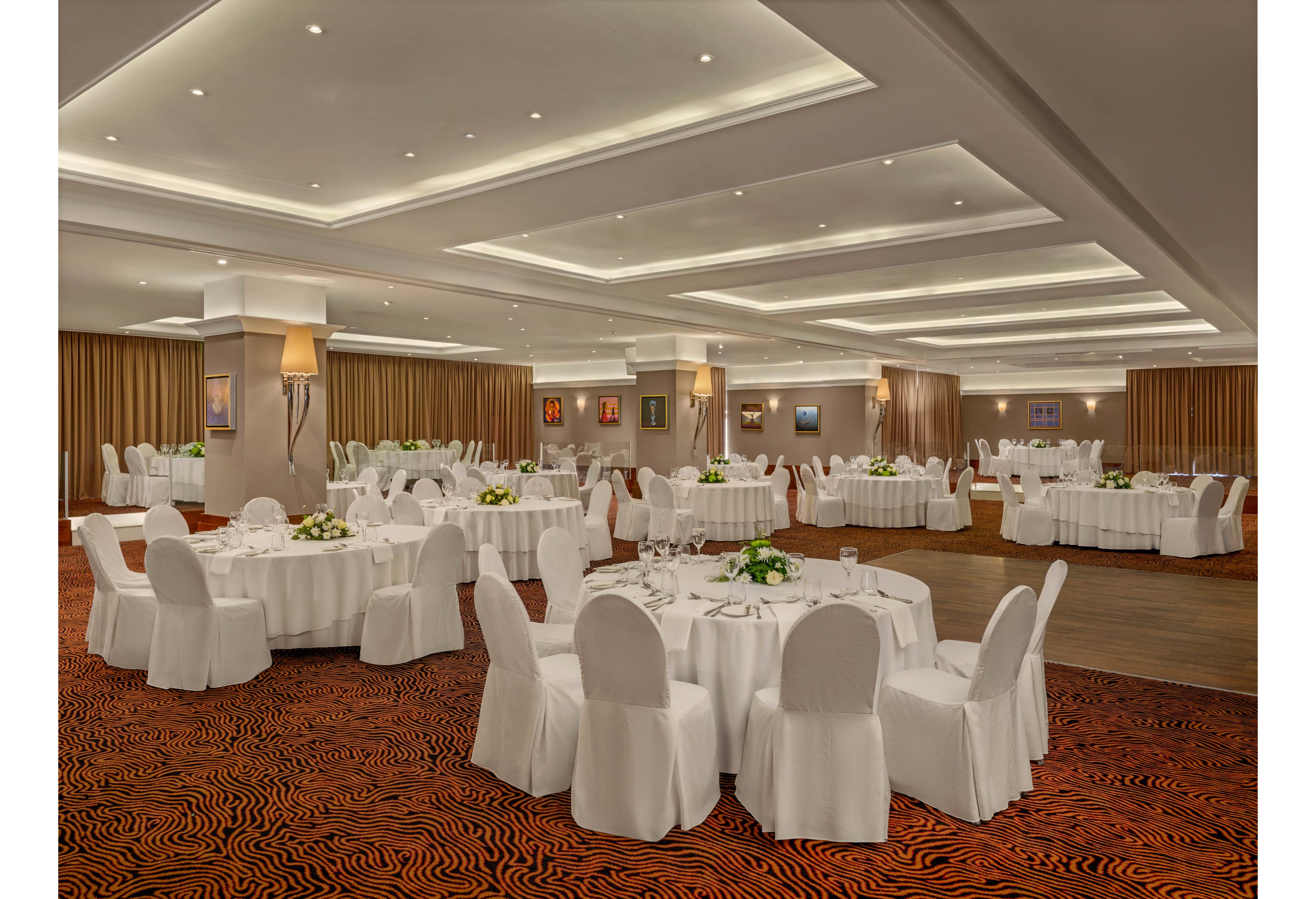 A spacious banquet hall is arranged with multiple round tables covered in white linens and matching chair covers. Each table is set with glassware and simple floral centerpieces, ready for a formal event or reception. The room features recessed ceiling lights, warm wall sconces, and framed artwork along the walls. A patterned carpet and floor-to-ceiling curtains add texture and warmth to the sophisticated interior setting.