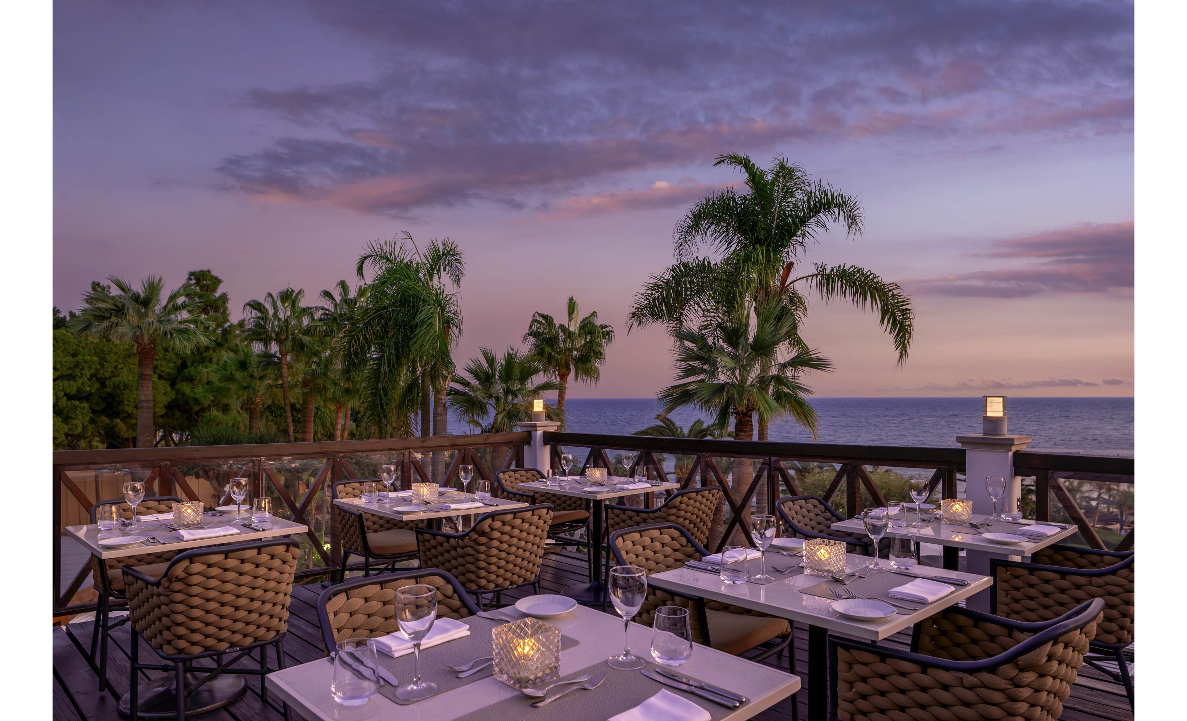 An elegant outdoor restaurant terrace overlooks the sea at dusk. Neatly set tables with white linens, glassware, and candlelit lanterns create a romantic dining atmosphere. Palm trees frame the view, and the sky glows with soft purple and pink tones over the water. The scene is calm and inviting, suggesting an upscale coastal dining experience.