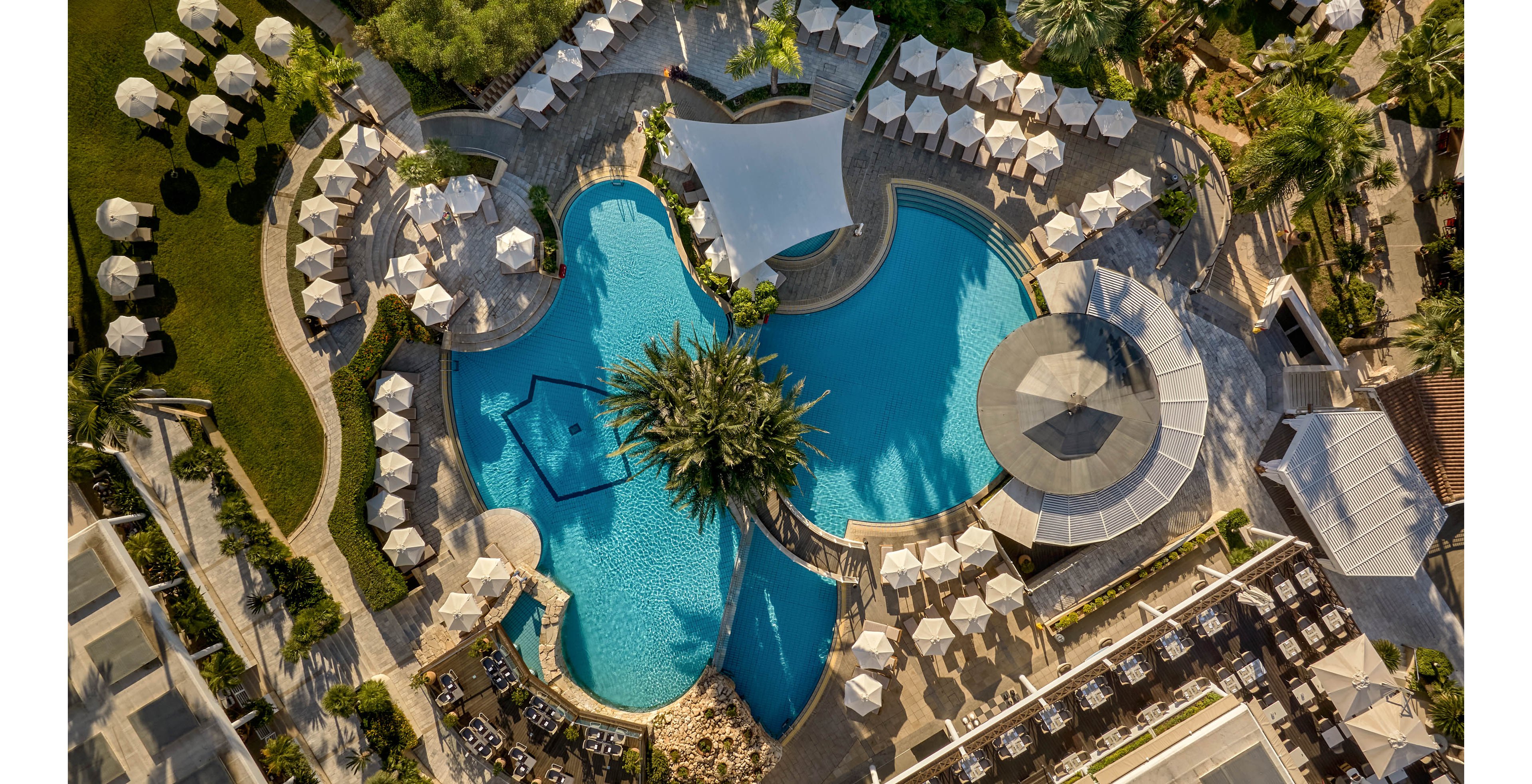 An aerial view showcases a large resort swimming pool surrounded by sun loungers and umbrellas. The pool curves organically around a central palm tree island, creating a tropical oasis feel. White parasols, green lawns, and landscaped pathways frame the bright blue water. Buildings, cabanas, and shaded seating areas indicate a luxurious vacation setting. The warm sunlight enhances the inviting atmosphere of relaxation and leisure.