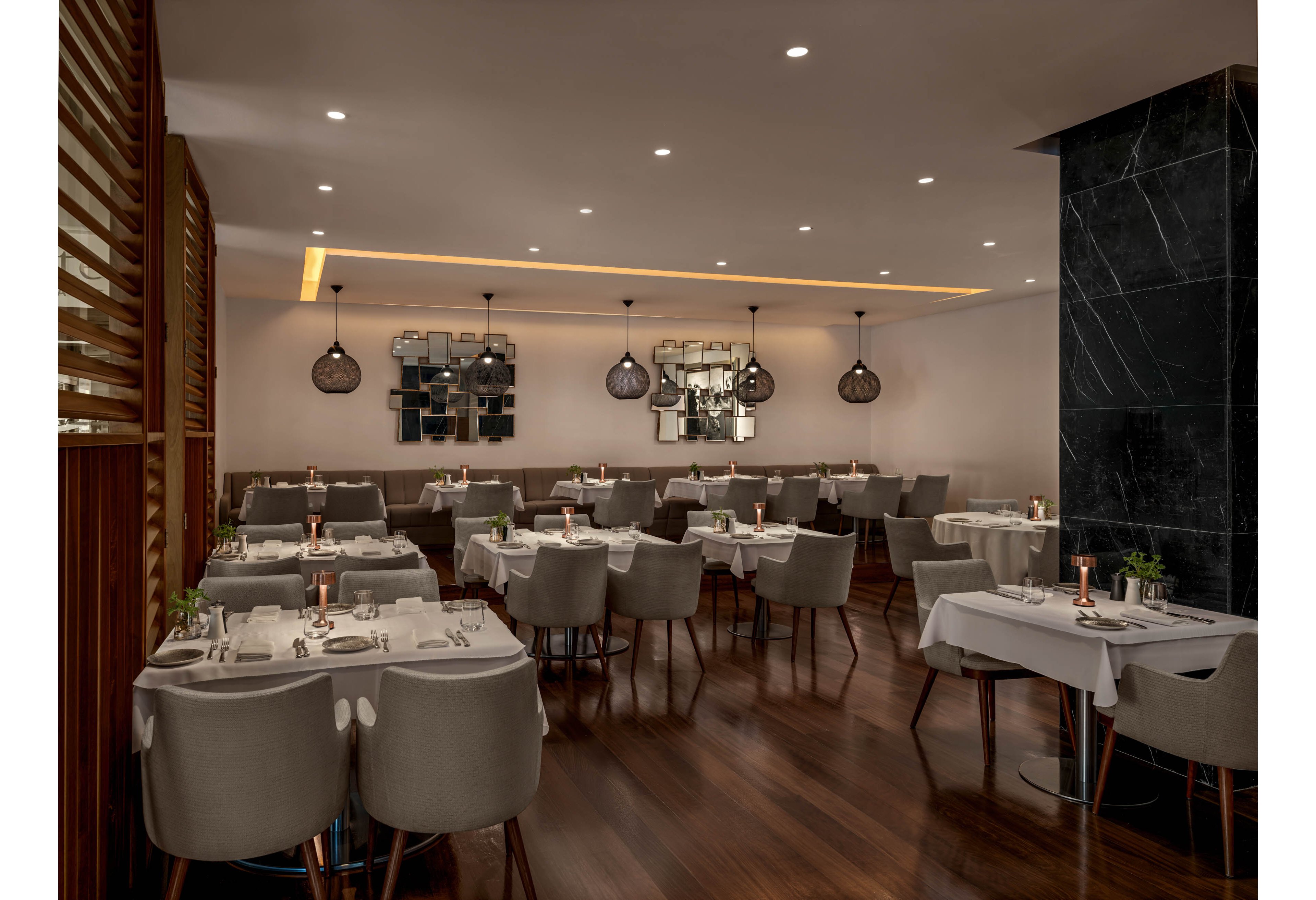 A contemporary restaurant dining room features neatly arranged tables with white tablecloths and cushioned gray chairs. Warm pendant lights and recessed ceiling lighting create a cozy, intimate atmosphere. Copper-colored candle holders and small green plants decorate each table, adding subtle elegance. Dark wood flooring and a black marble column contrast with the soft neutral tones of the seating and walls.
